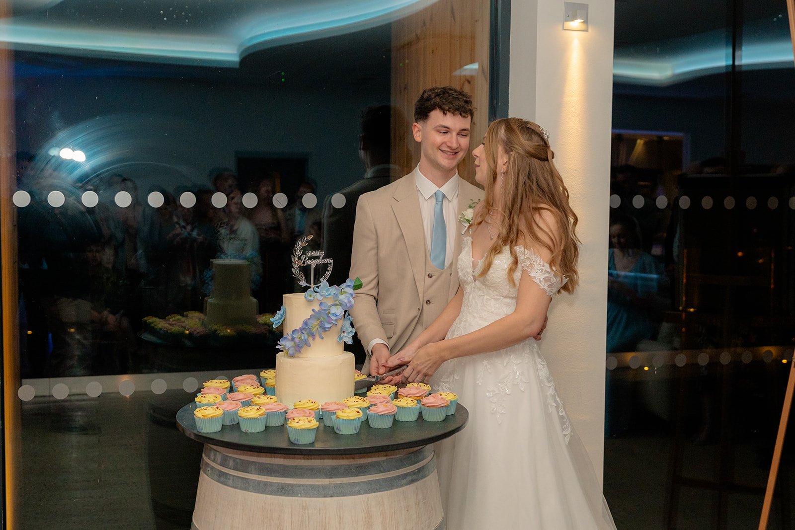 Hannah & Cameron cut their wedding cake together during their evening reception at Sandy Cove Hotel in North Devon. The couple smile beside a decorated cake and cupcakes while guests watch through the window reflections behind them.