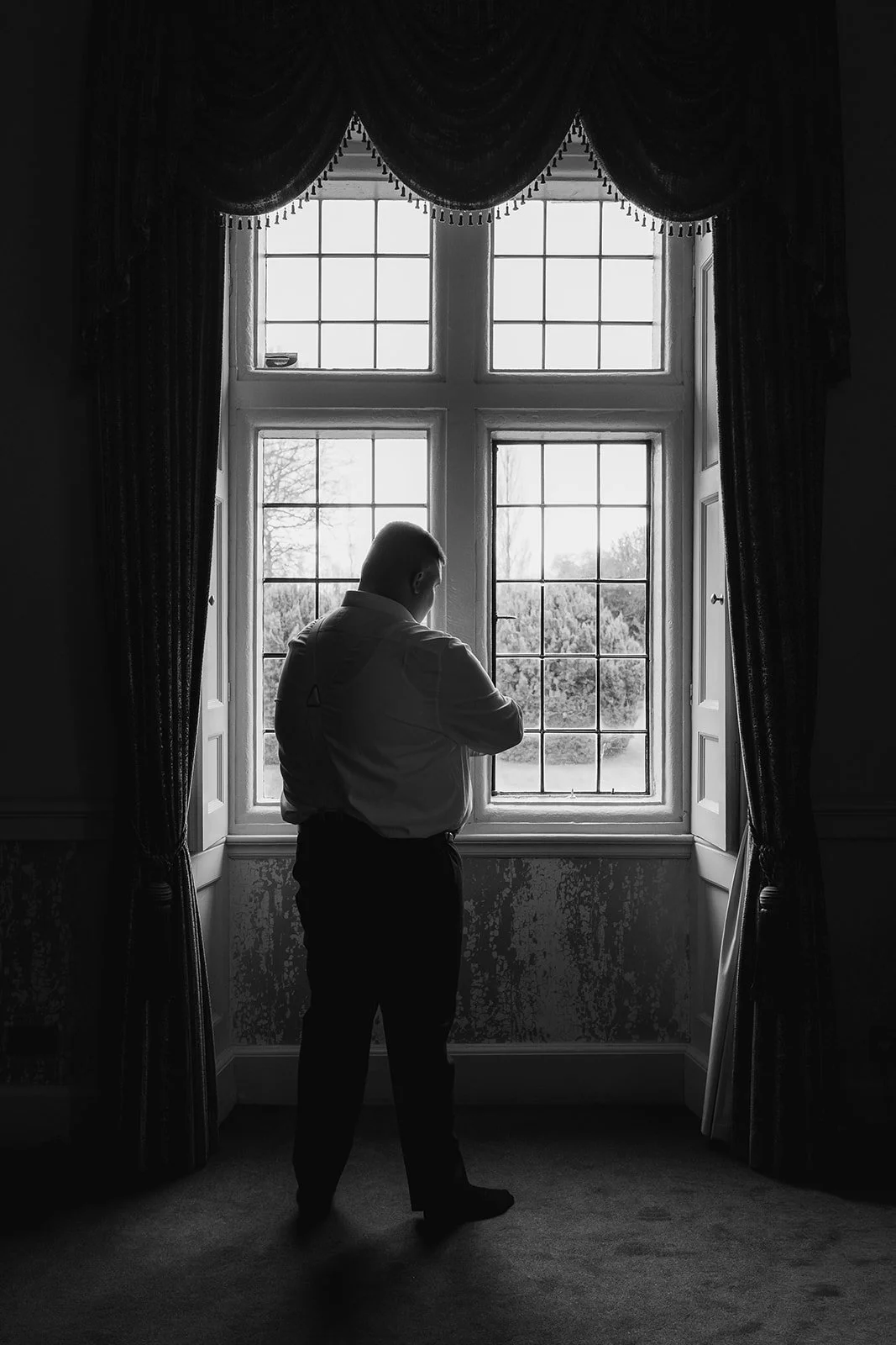 Rob stands in silhouette by a leaded window, adjusting his shirt during Becky & Rob’s wedding at Clearwell Castle in Gloucestershire, UK. Black and white groom prep in the castle suite, Clearwell Castle wedding photography. Soft window light.