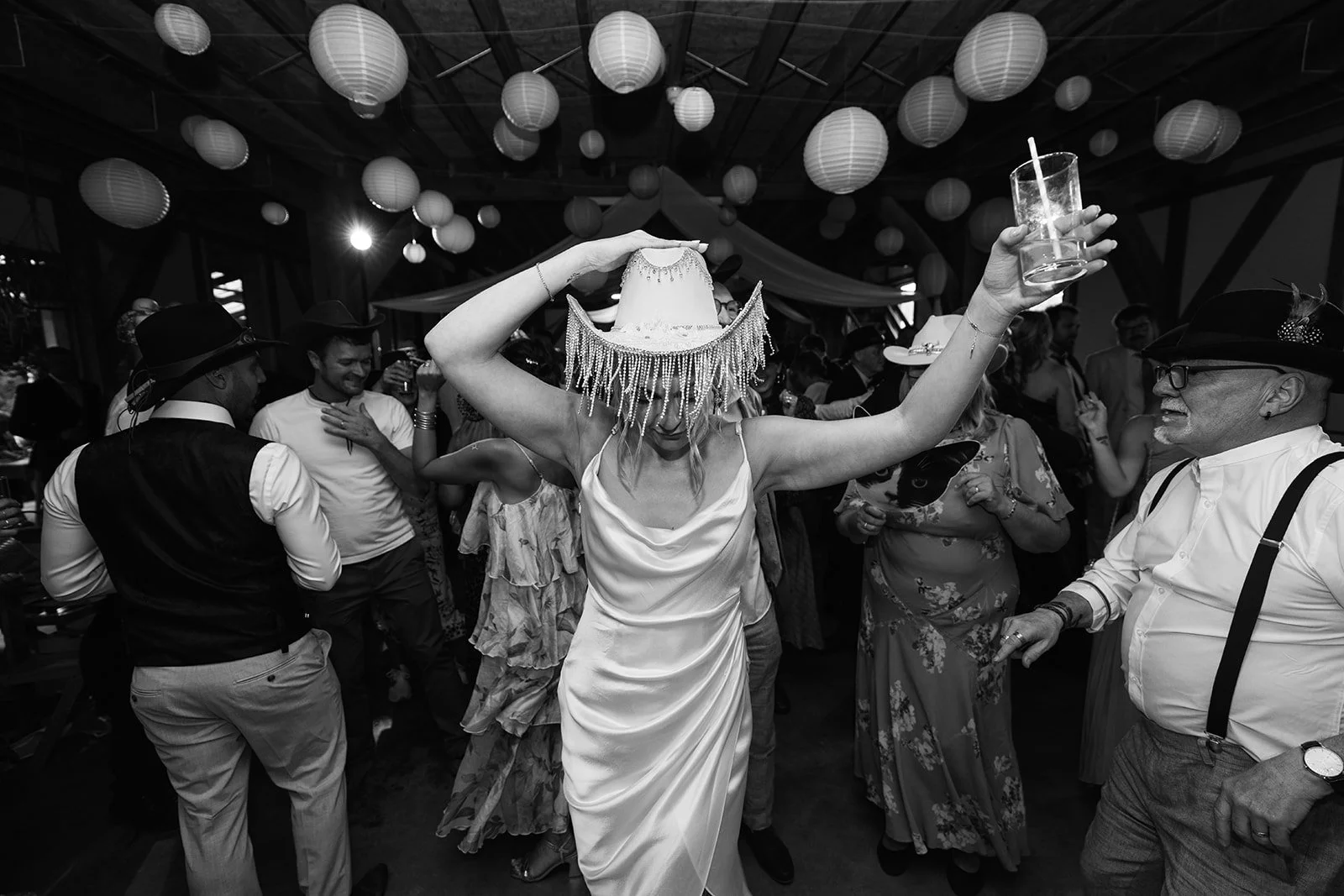 Black and white dance floor photo from Hannah & Ben’s wedding at Grays Barn in Devon, UK, with Hannah dancing in a fringed cowboy hat, drink raised as guests celebrate beneath lanterns, captured with lively party energy by Just James Weddings.
