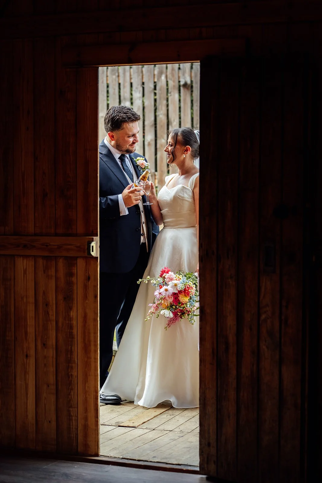 Harriet & Simon smile at each other while holding champagne and Harriet’s colourful bouquet in a barn doorway at Dovecote Events in Oxfordshire, photographed by Just James Weddings.