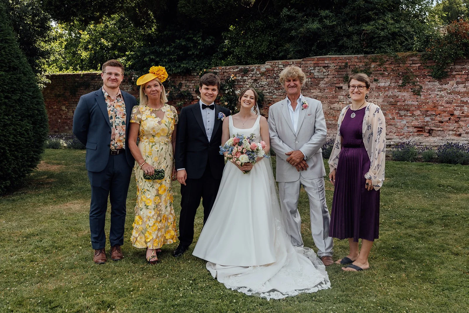 Rachel & Tom pose with four wedding guests in the gardens at Brympton House in Somerset, with a red brick wall behind the group, photographed by Just James Weddings.
