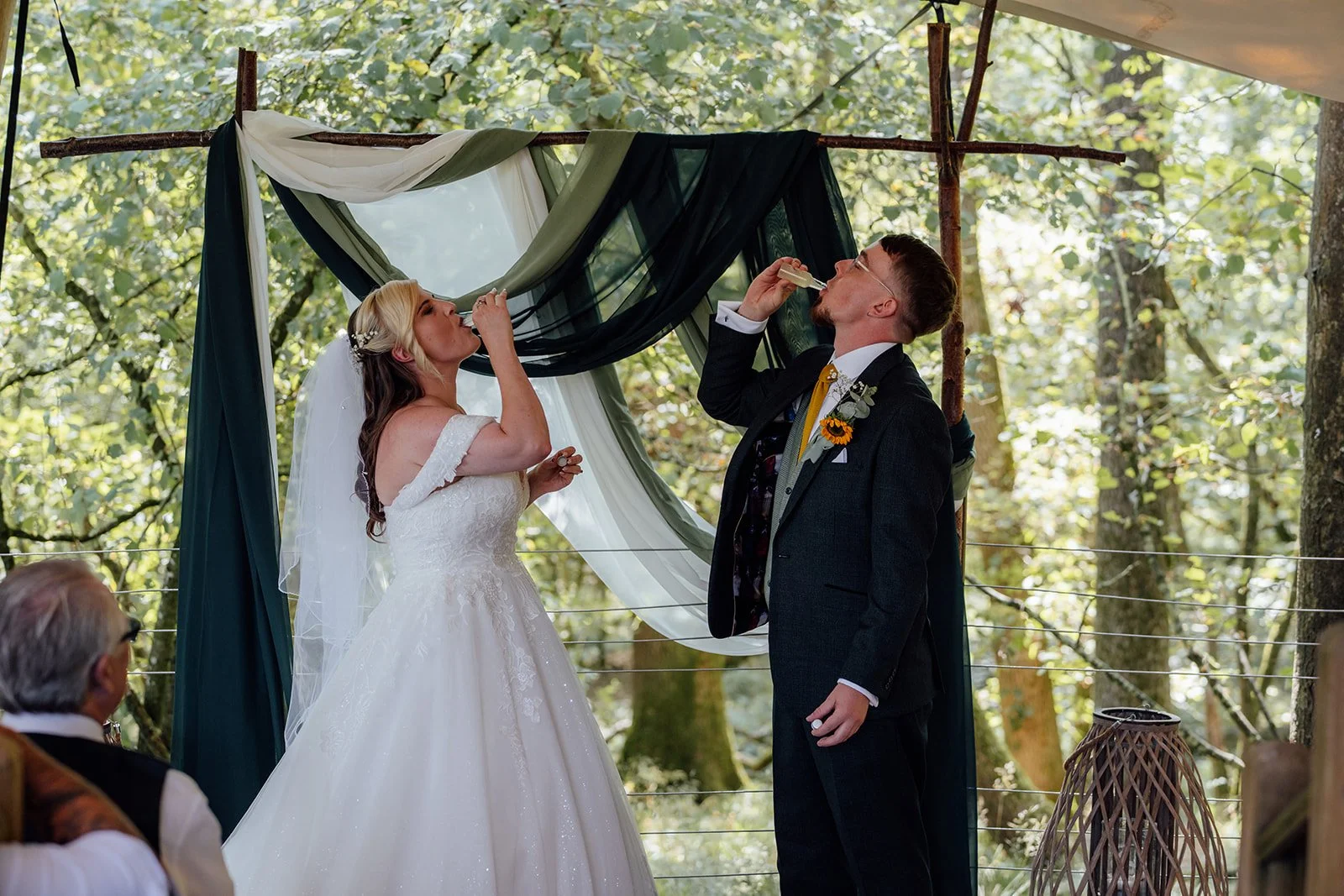Lucy and Matthew sharing a playful drink during their woodland wedding ceremony at Curated Canvas at Noah’s Pond in Somerset, photographed by Just James Weddings.