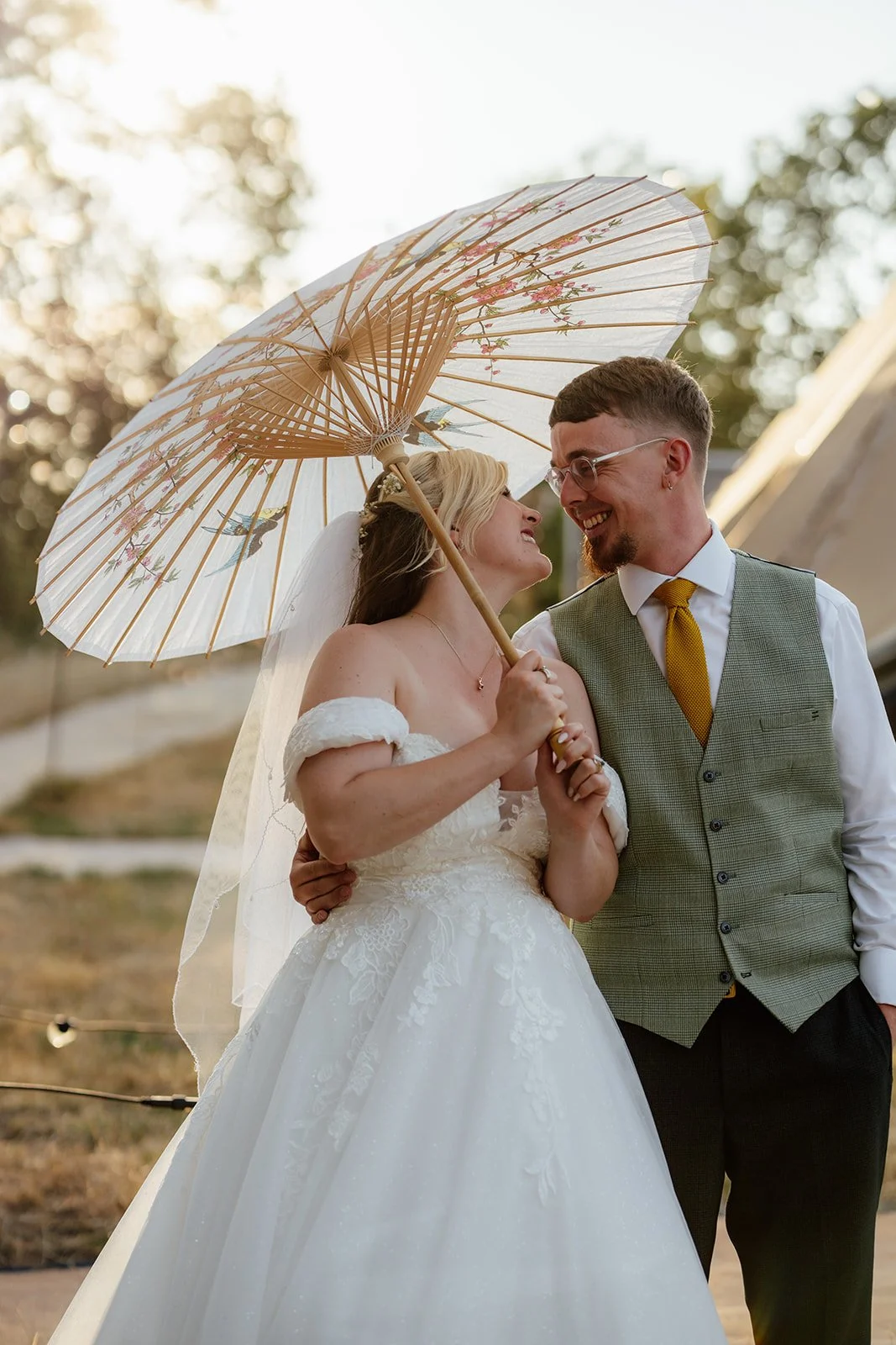 Lucy and Matthew smiling together beneath a decorative parasol during golden hour wedding portraits at Curated Canvas at Noah’s Pond in Somerset, photographed by Just James Weddings.