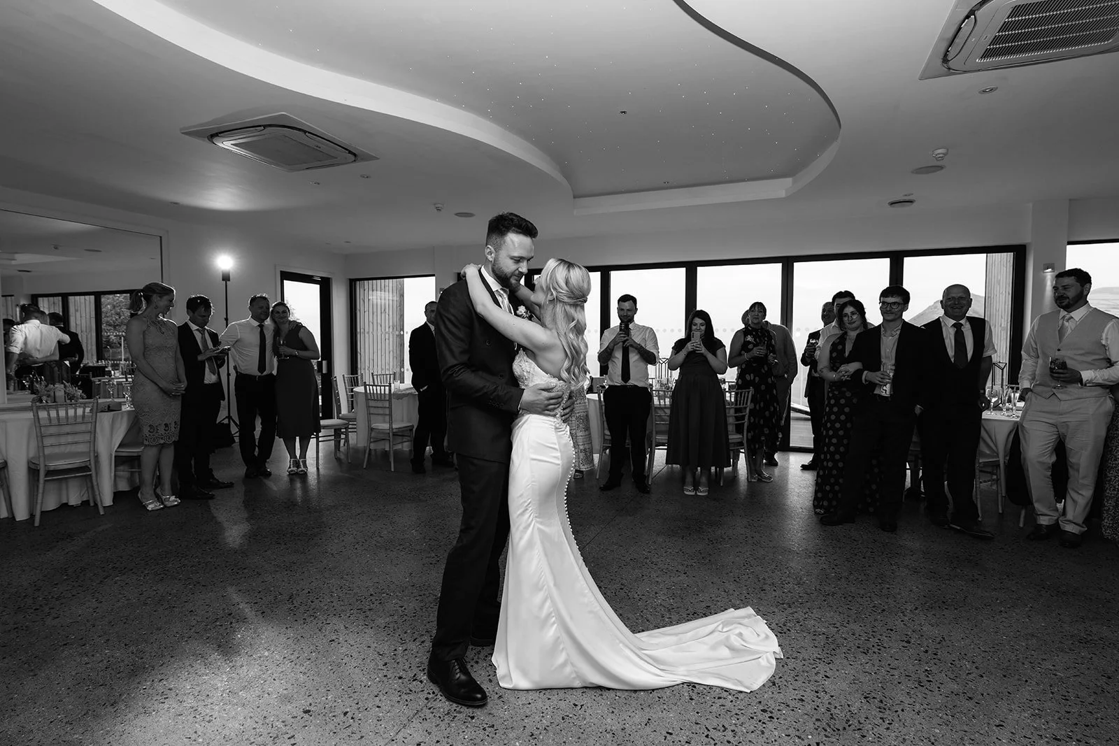 Black-and-white photo of Amy & Connor sharing their first dance as wedding guests watch inside the reception room at Sandy Cove Hotel in North Devon, UK.