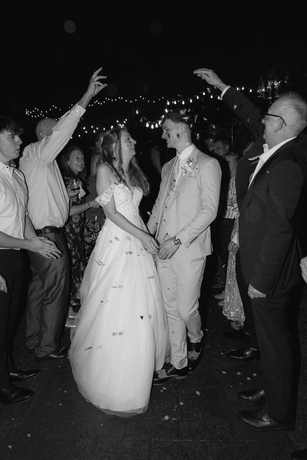 Hannah & Cameron walk through a joyful confetti tunnel created by guests during their evening celebration at Sandy Cove Hotel, North Devon. Friends and family cheer beneath festoon lights as the newlyweds laugh together.