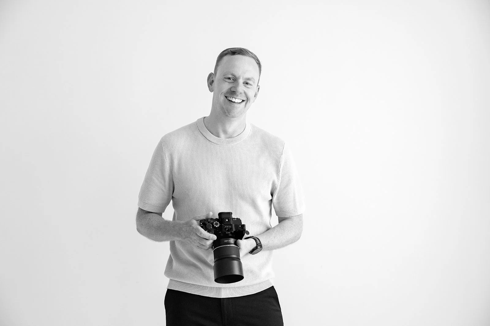 Black and white portrait of James from Just James Weddings smiling while holding his camera in a clean studio setting, for his Somerset wedding photography website profile.