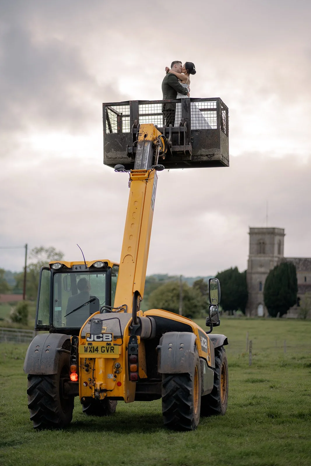 Jaye & Josh kiss high above Farrington's Farm in Bristol on a JCB lift, with countryside fields, a distant church and soft cloudy skies creating a playful wedding portrait full of personality, joy and brilliant farm celebration charm together.