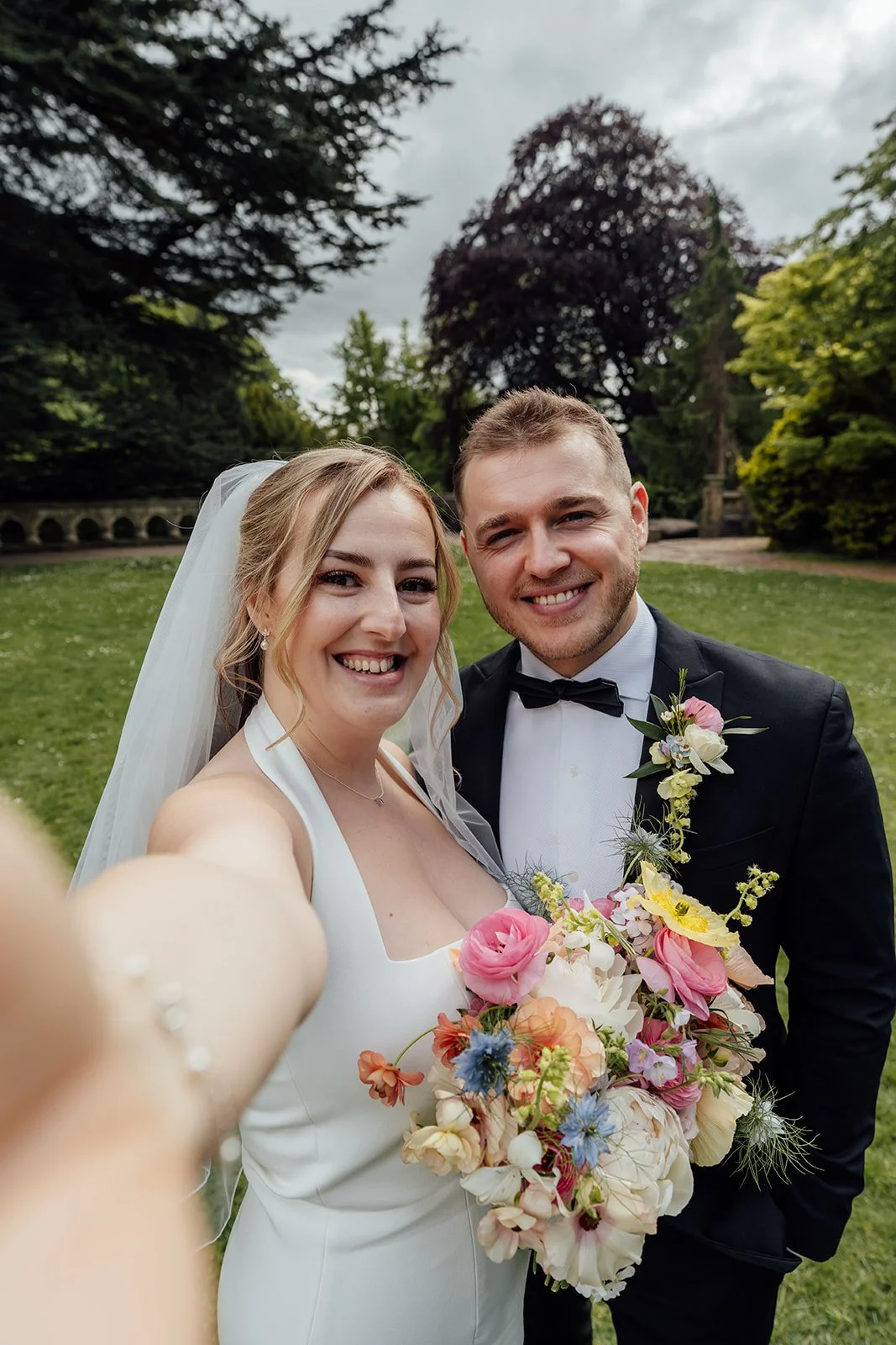 Bryony & Max smile for a close-up selfie-style portrait at Insole Court in Cardiff, with Bryony holding her colourful bouquet, Max in a black suit and the venue’s green lawn and trees creating a relaxed wedding backdrop around them on the lawn.