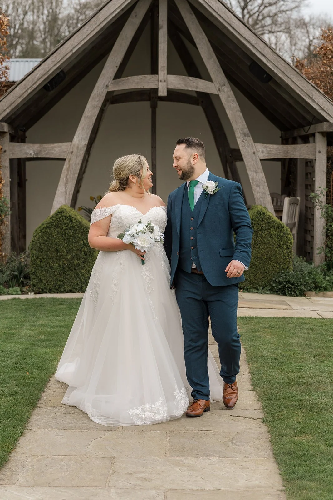 Siobhan & Nicholas walk together along the garden path at Kingscote Barn in the Cotswolds, UK, smiling at each other outside the rustic barn wedding venue, a relaxed countryside wedding portrait with wooden pavilion and bridal bouquet in view.