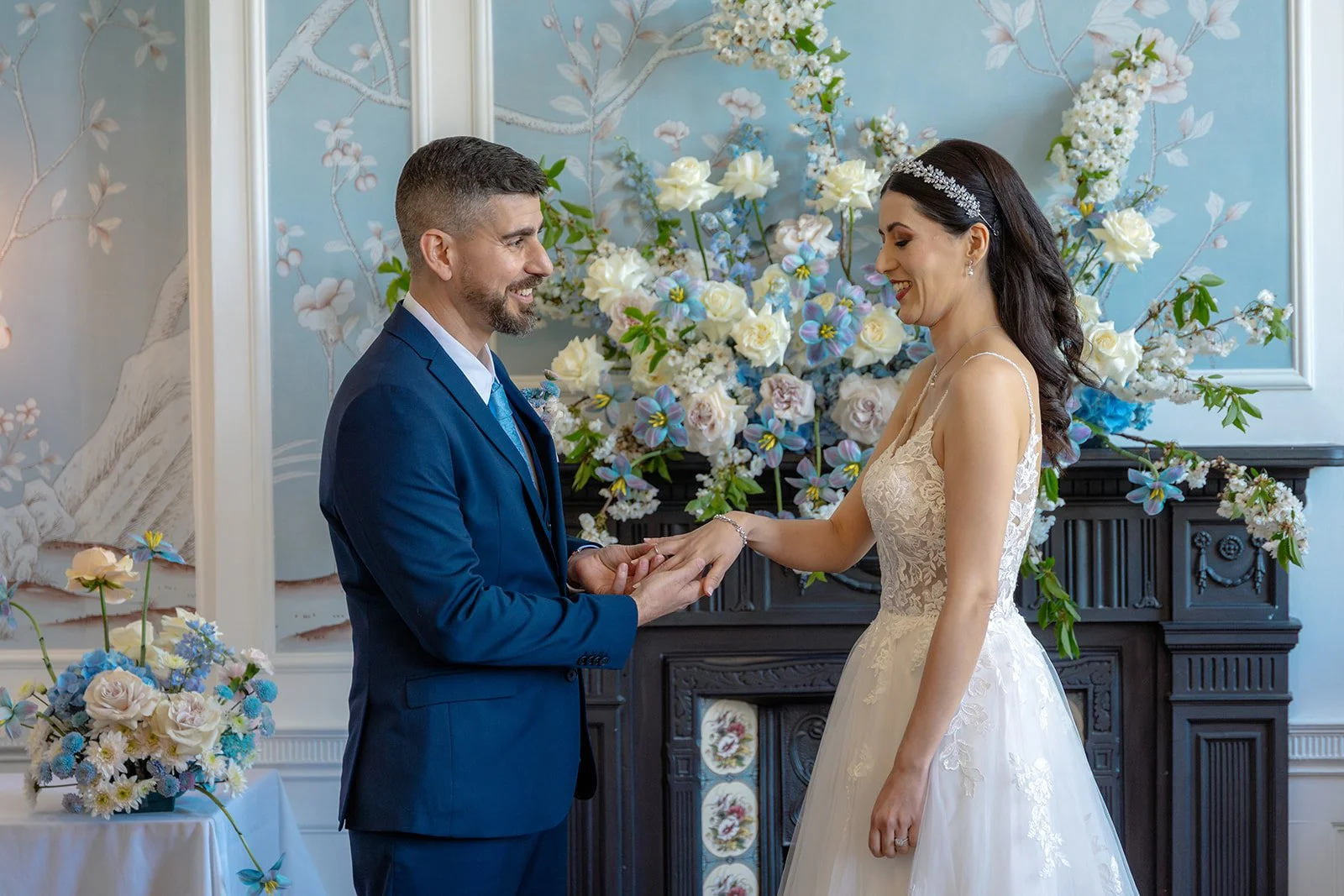 Ruth and Carmelo exchange rings during their wedding ceremony at No. 4 Clifton Village in Bristol, UK, surrounded by elegant pastel flowers and the venue’s distinctive blue décor.