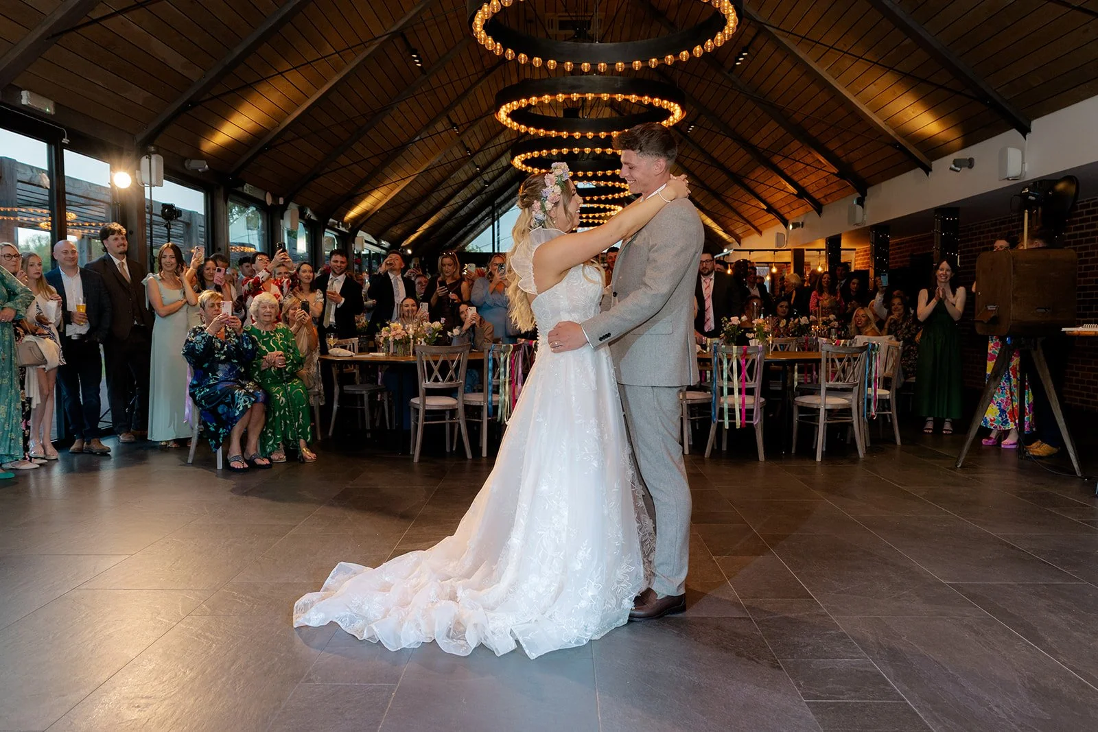 Jaye & Josh sharing their first dance inside Syrencot in Wiltshire, surrounded by guests and warm circular pendant lights, photographed by Just James Weddings.