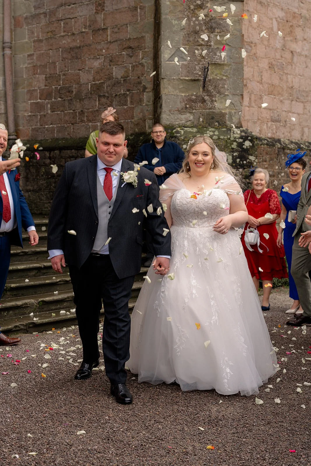 Becky & Rob walk hand in hand through confetti outside Clearwell Castle in Gloucestershire, UK after their ceremony. Guests celebrate as petals fall around the couple during their joyful Clearwell Castle wedding confetti moment.