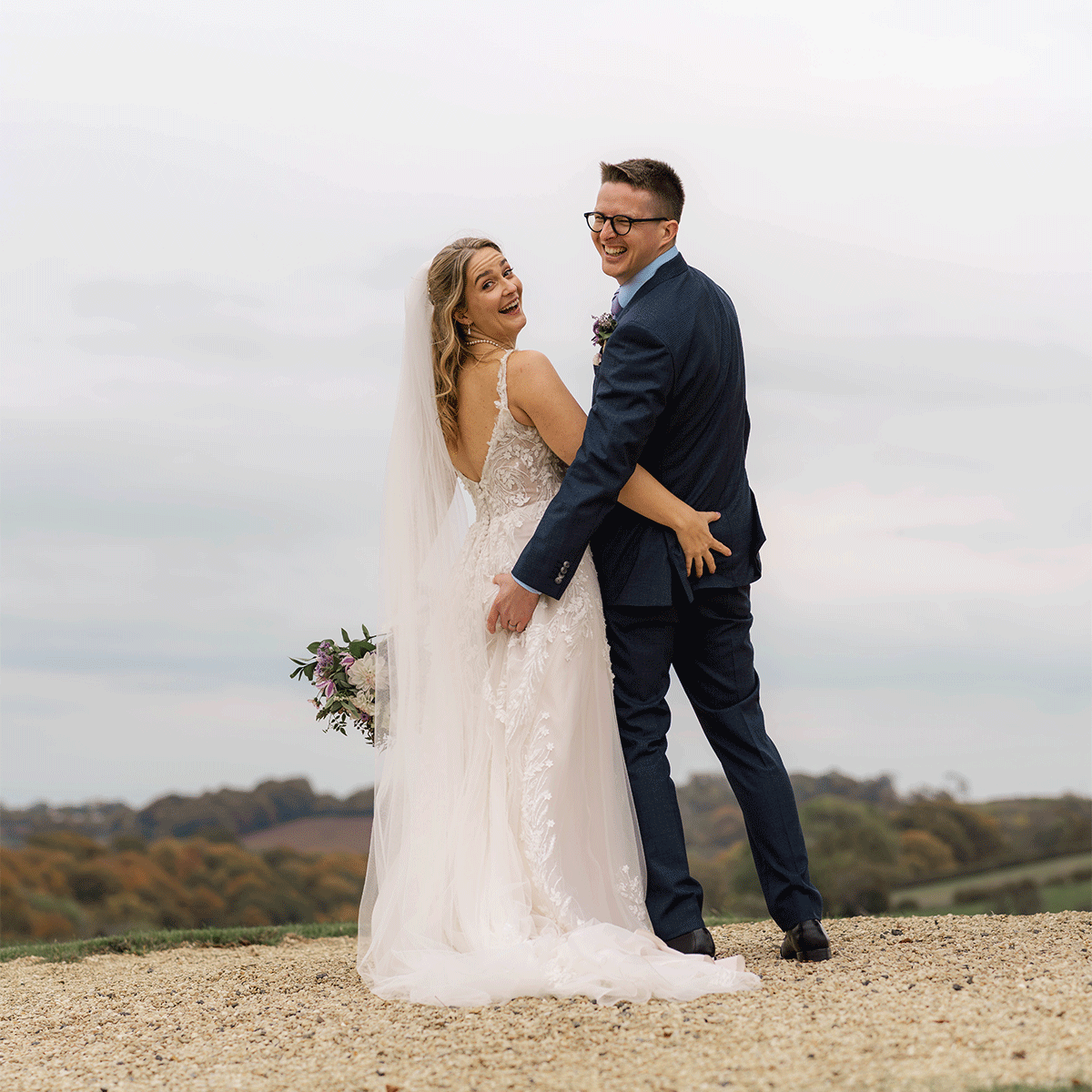 Izzy & Andy standing on a hilltop on their wedding day at The Kingscote Barn, cotswolds. The bride looks back and smiles over her shoulder while holding her bouquet as the groom laughs beside her, with rolling countryside and pale sky.