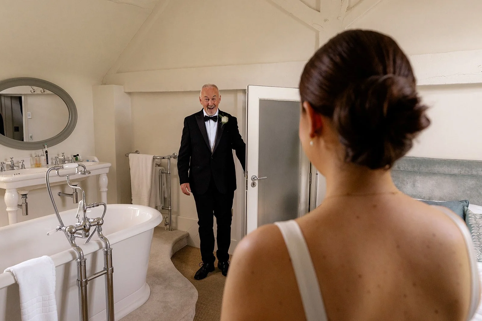 Lydia seen from behind as she has a quiet first look with a suited family member in a bright room at Hencote Estate in Shropshire, UK, during Lydia & Kieran's wedding morning, beside a freestanding bath, soft neutral decor and warm indoor light.
