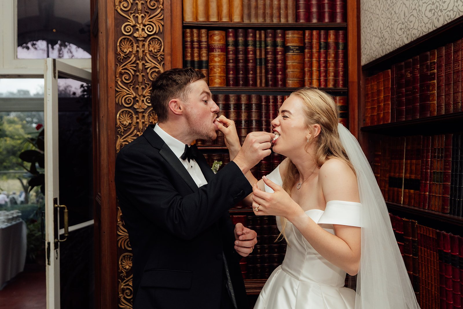 Caitlin and Kevin laughing as they feed each other sweets during their wedding reception at Clevedon Hall in Somerset, photographed in front of the venue’s traditional library shelves.