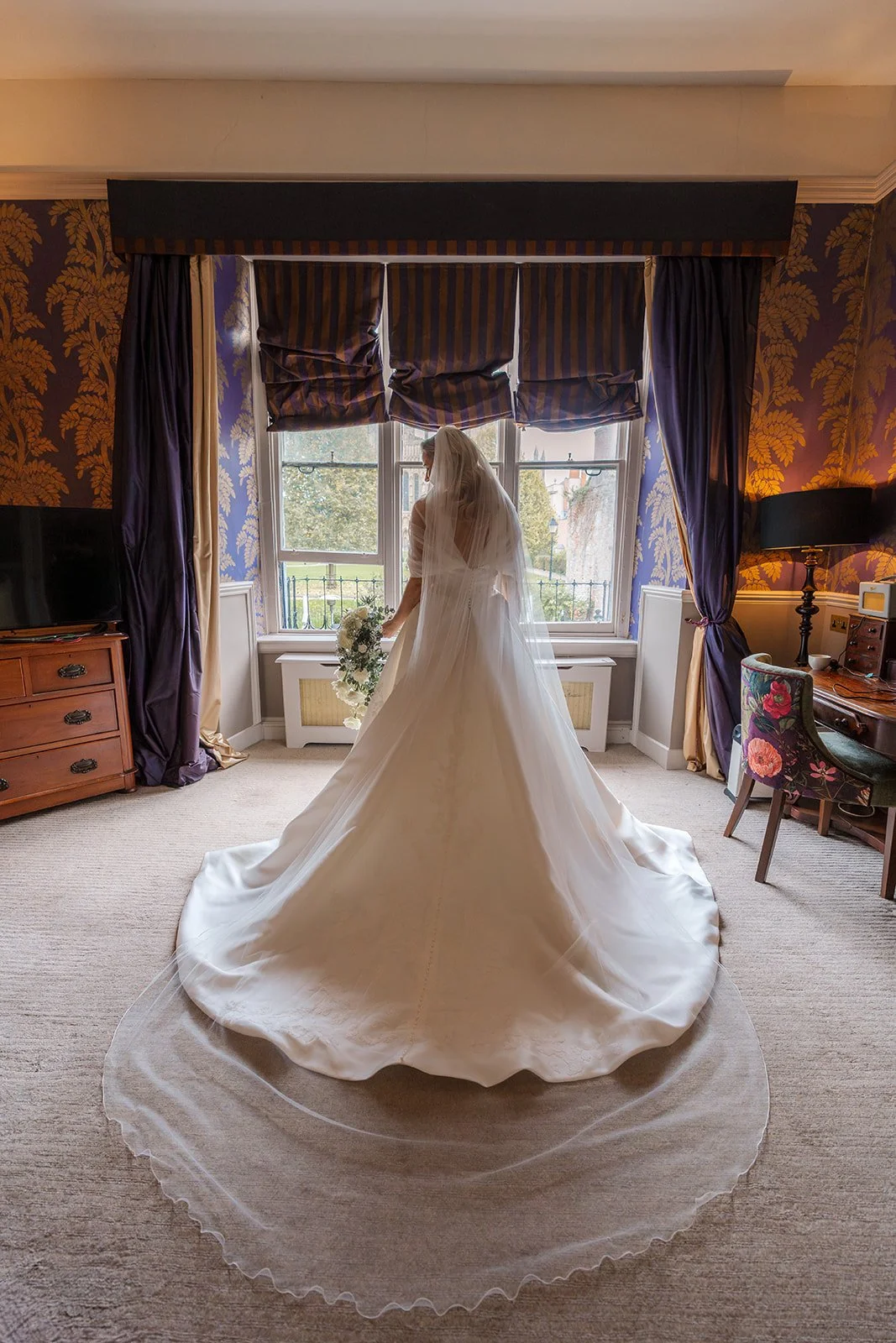 Iona stands by the window in her bridal suite at Bishop’s Palace in Somerset, UK, wearing a flowing wedding dress and veil with a long train spread across the carpet, framed by elegant interiors in a timeless bride getting ready.