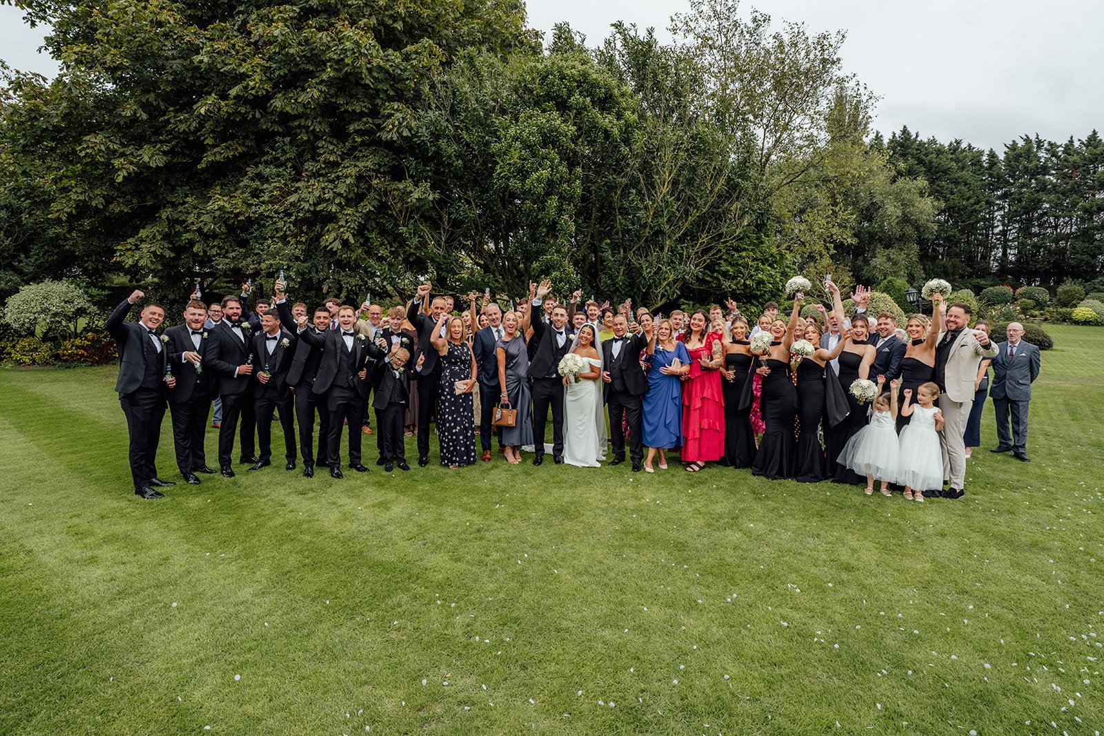 Nisha and Jack stand with their wedding guests on the gardens at Batch Country House in Somerset, UK, with family and friends raising their hands and celebrating around the newlyweds after the ceremony, photographed by Just James Weddings.