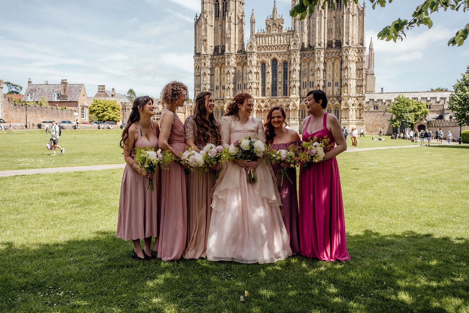 Charlotte & Jack wedding party portrait at Bishop’s Palace in Somerset, UK, with the bride and bridesmaids in pink and magenta dresses laughing together on the lawn, holding bouquets as Wells Cathedral stands behind them in warm summer light.