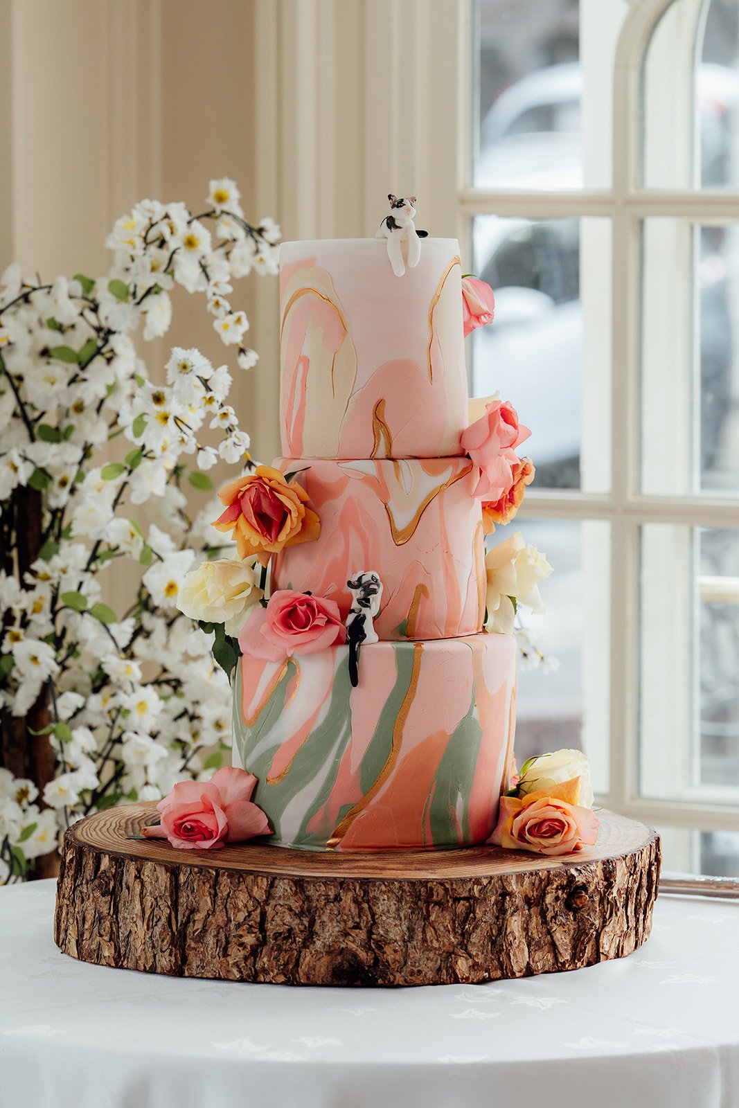 Kasey and Larisha’s three-tier wedding cake decorated with pink, peach and cream details, roses and playful cat toppers at Oaks Farm Weddings in Surrey.