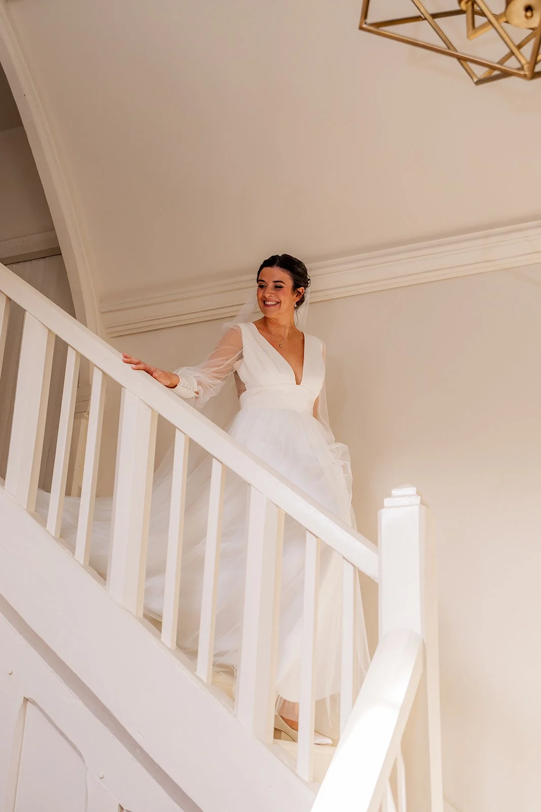 Megan descends a white staircase at The Great Barn Devon during Megan & Luca’s wedding, smiling softly as her flowing gown and veil catch the warm light. The clean interior lines and chandelier above frame this elegant Devon bridal moment.