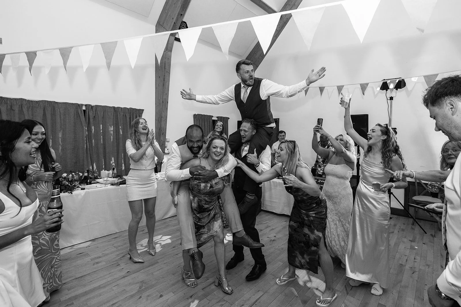 Black-and-white candid of Aaron & Jack being lifted onto guests’ shoulders as friends dance and cheer during their lively evening reception at Rock Village Hall in Worcestershire, photographed by Just James Weddings.