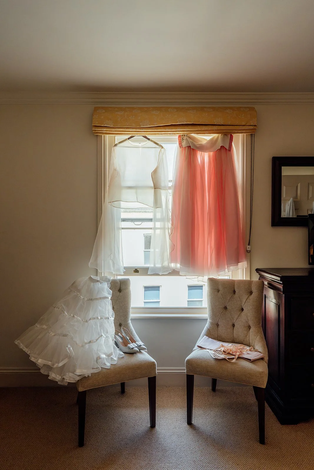 Charlotte & Jack wedding details at Bishop’s Palace in Somerset, UK, with sheer bridalwear and a pink dress hanging in a window above chairs, shoes and accessories in a softly lit preparation room before their ceremony in Wells.