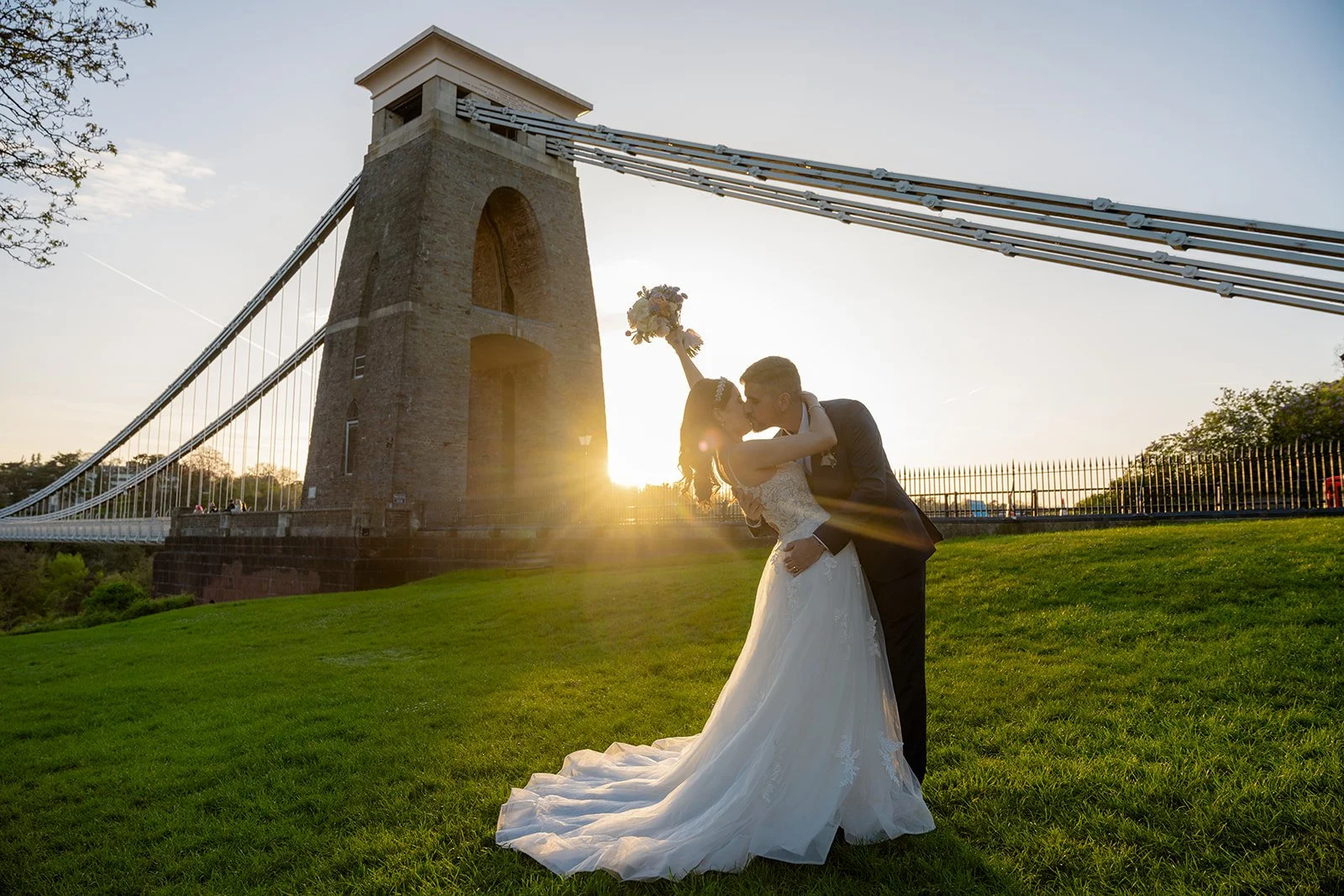 Ruth and Carmelo share a romantic kiss at sunset with Clifton Suspension Bridge behind them in Bristol, UK, creating a scenic wedding portrait near No. 4 Clifton Village.