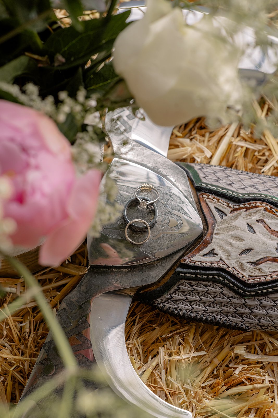 Jaye & Josh's Farrington's Farm wedding in Bristol with their rings resting on a decorated double axe among straw, soft greenery and blurred pink and white flowers, tying their playful farm celebration to personal handmade wedding details too.