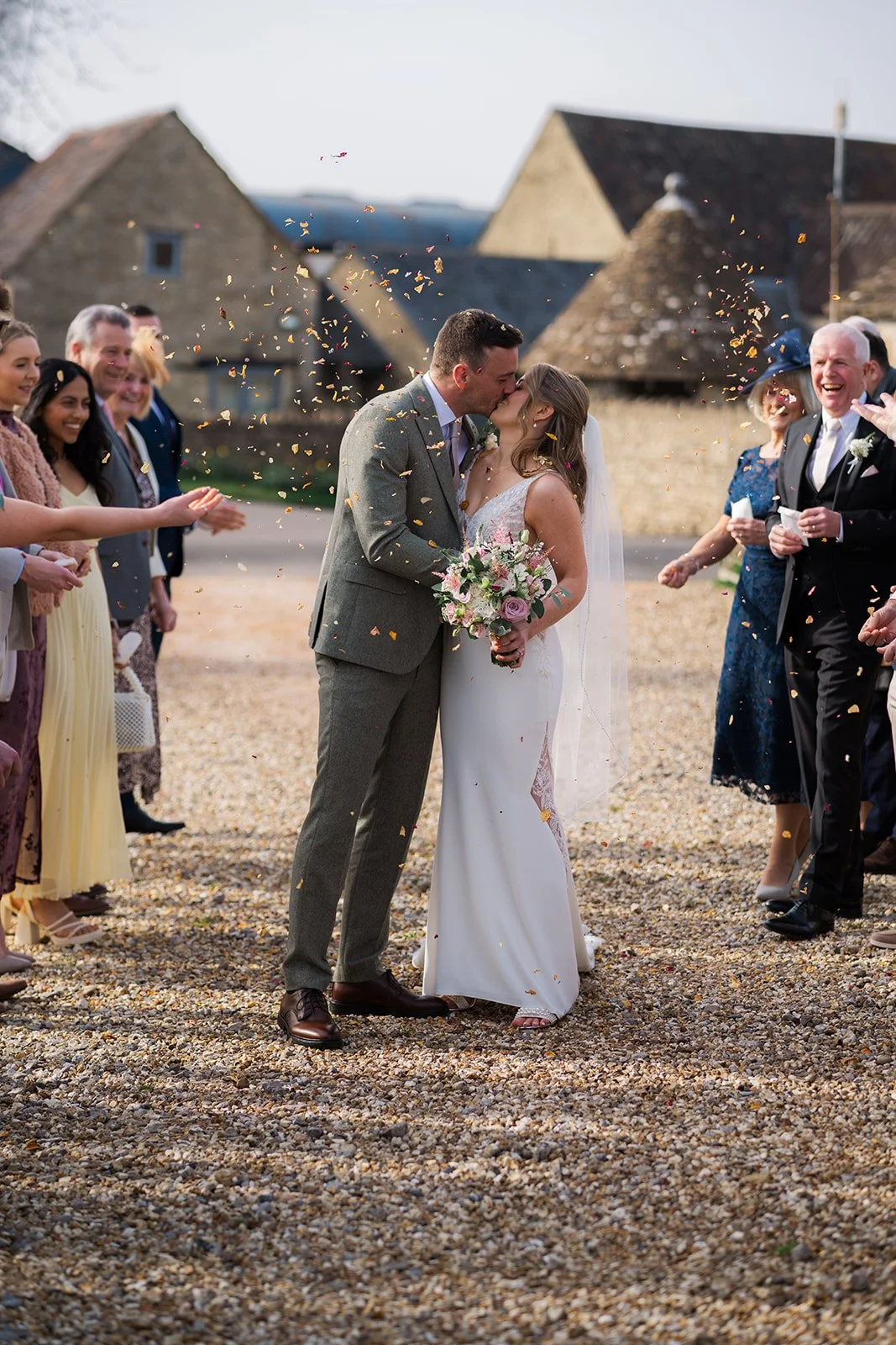 Anna and Tom kiss during their confetti exit at Winkworth Farm in the Cotswolds, UK, surrounded by cheering guests and falling petals. This outdoor wedding photo captures a joyful just-married moment in beautiful Cotswolds wedding photography.