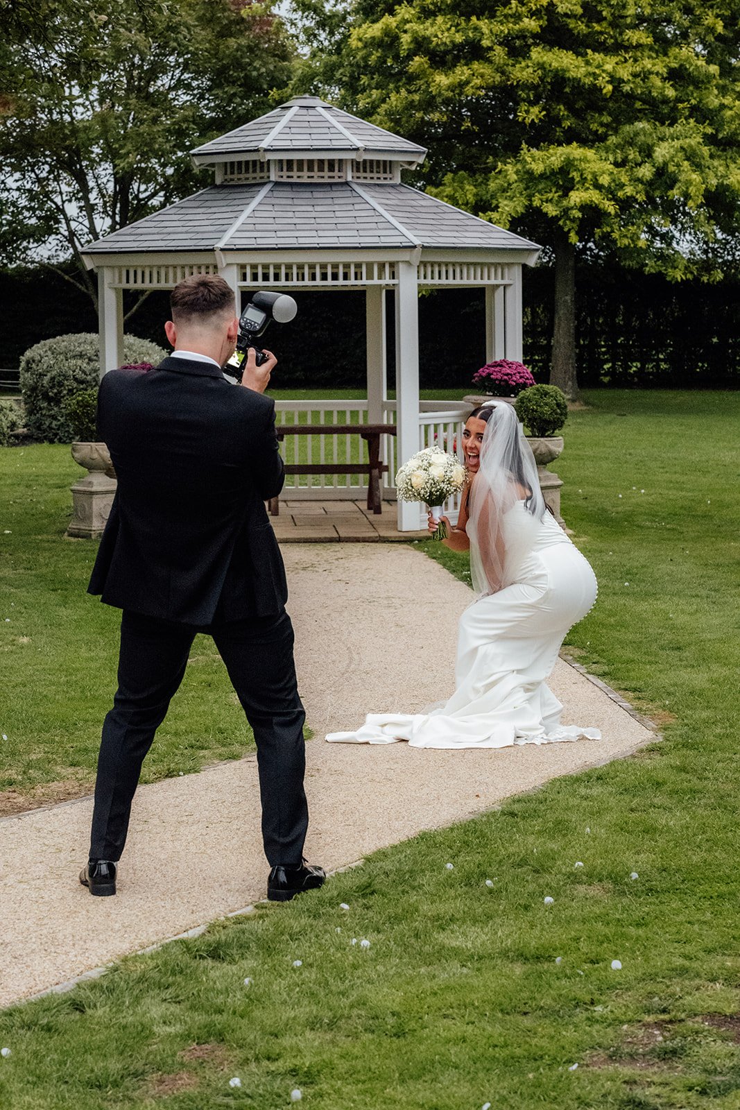 Nisha laughs while posing with her bouquet as Jack photographs her in the gardens at Batch Country House in Somerset, UK, with a white gazebo and manicured lawn behind them, capturing a playful wedding moment photographed by Just James Weddings.