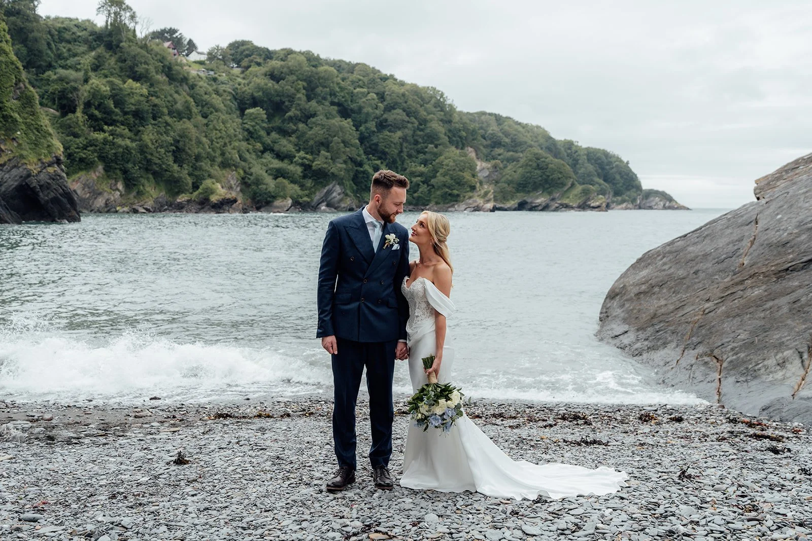Amy & Connor stand on a pebble beach at Sandy Cove Hotel in North Devon, UK, with the bride holding a blue and white bouquet as waves roll in beneath rocky, tree-covered cliffs.