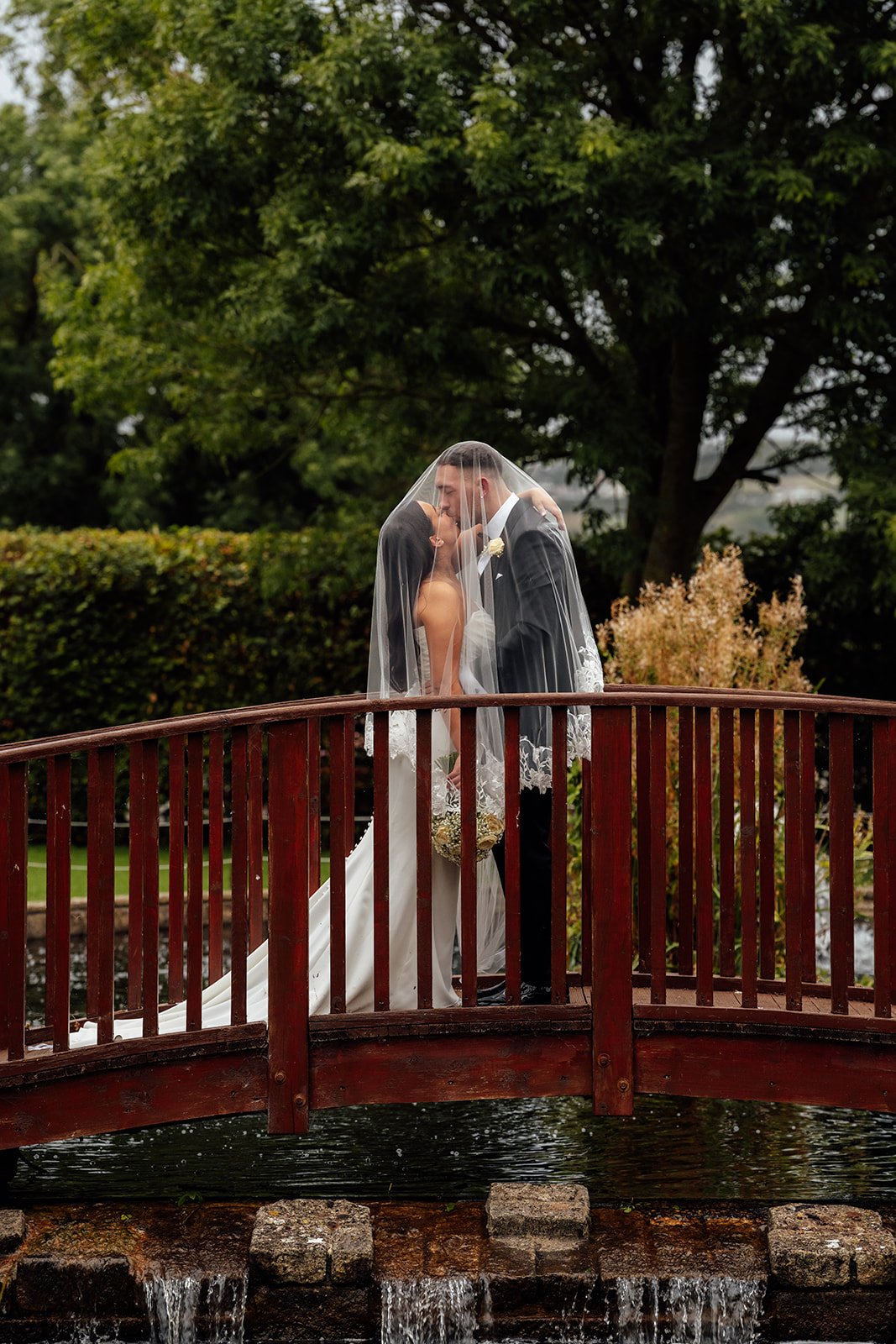 Nisha and Jack kiss beneath Nisha’s veil on a wooden bridge at Batch Country House in Somerset, UK, surrounded by lush greenery and water as her long white dress trails behind them in a romantic wedding portrait together by Just James Weddings.