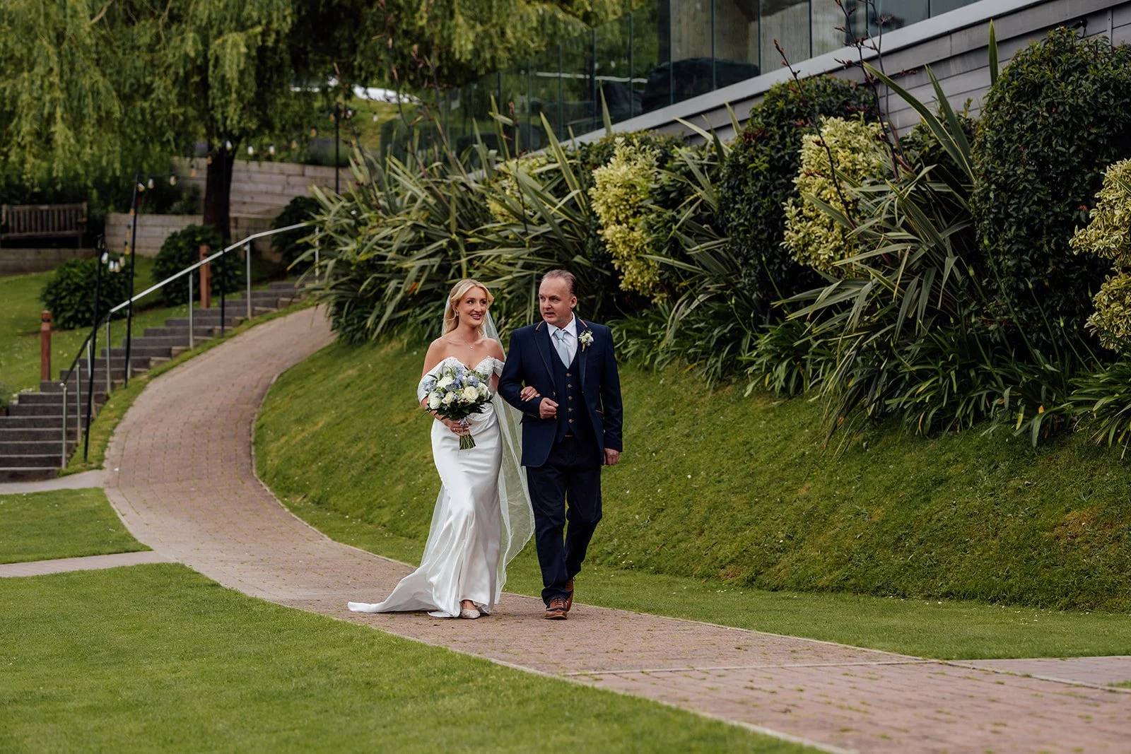 Amy walks arm in arm with her father through the gardens towards the outdoor wedding ceremony at Sandy Cove Hotel in North Devon, UK.