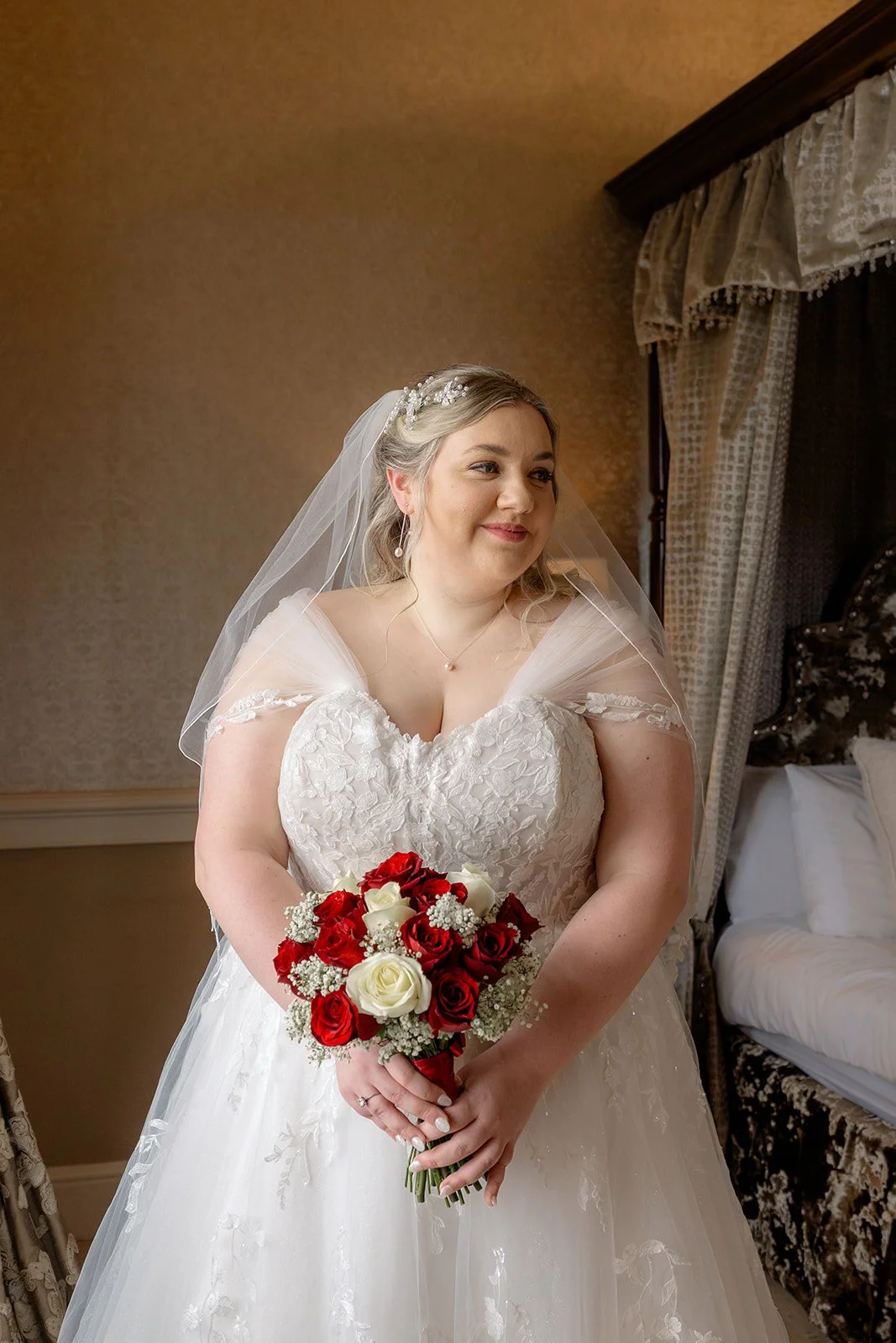 Becky holds a rose bouquet in her lace wedding dress, smiling in the bridal suite during Becky & Rob’s wedding at Clearwell Castle, Gloucestershire, UK. Bridal portrait for Clearwell Castle wedding photography and Gloucestershire wedding venue.