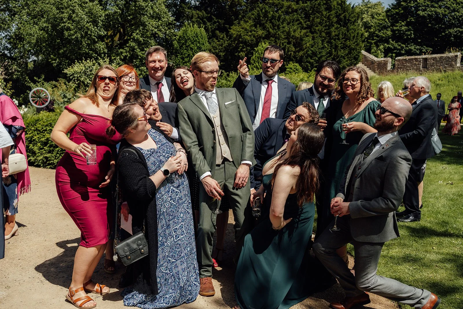 Charlotte & Jack with wedding guests at Bishop’s Palace in Somerset, UK, posing playfully in the gardens during sunny reception drinks, with friends laughing, holding glasses and crowding around the groom for a lively group portrait together.
