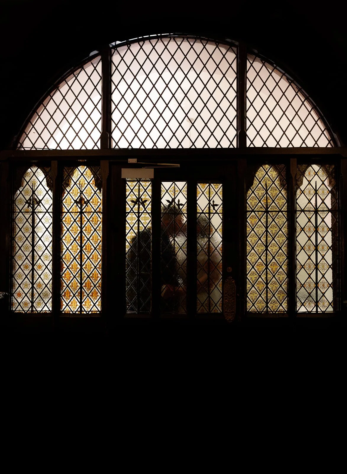 Becky & Rob share a quiet kiss behind the ornate glass doors at Clearwell Castle in Gloucestershire, UK. Silhouetted evening portrait of the couple through patterned windows during their romantic Clearwell Castle wedding night.