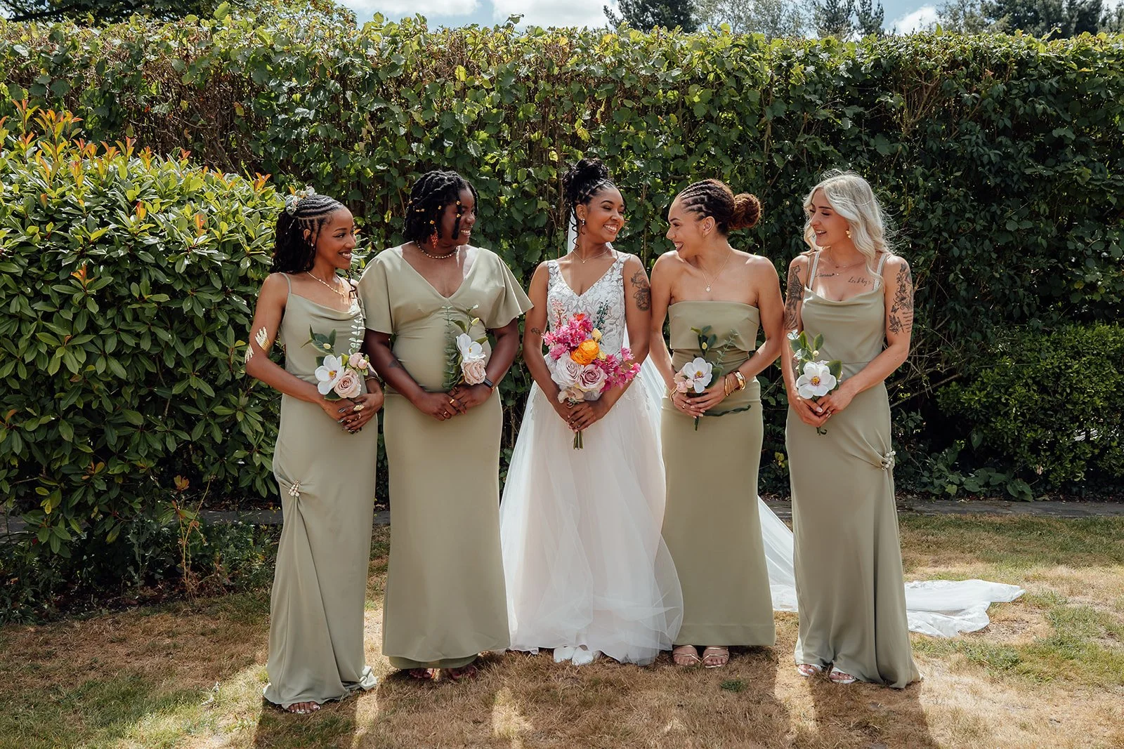 Larisha smiling with her bridesmaids in sage green dresses and holding colourful bouquets in the gardens at Oaks Farm Weddings in Surrey.