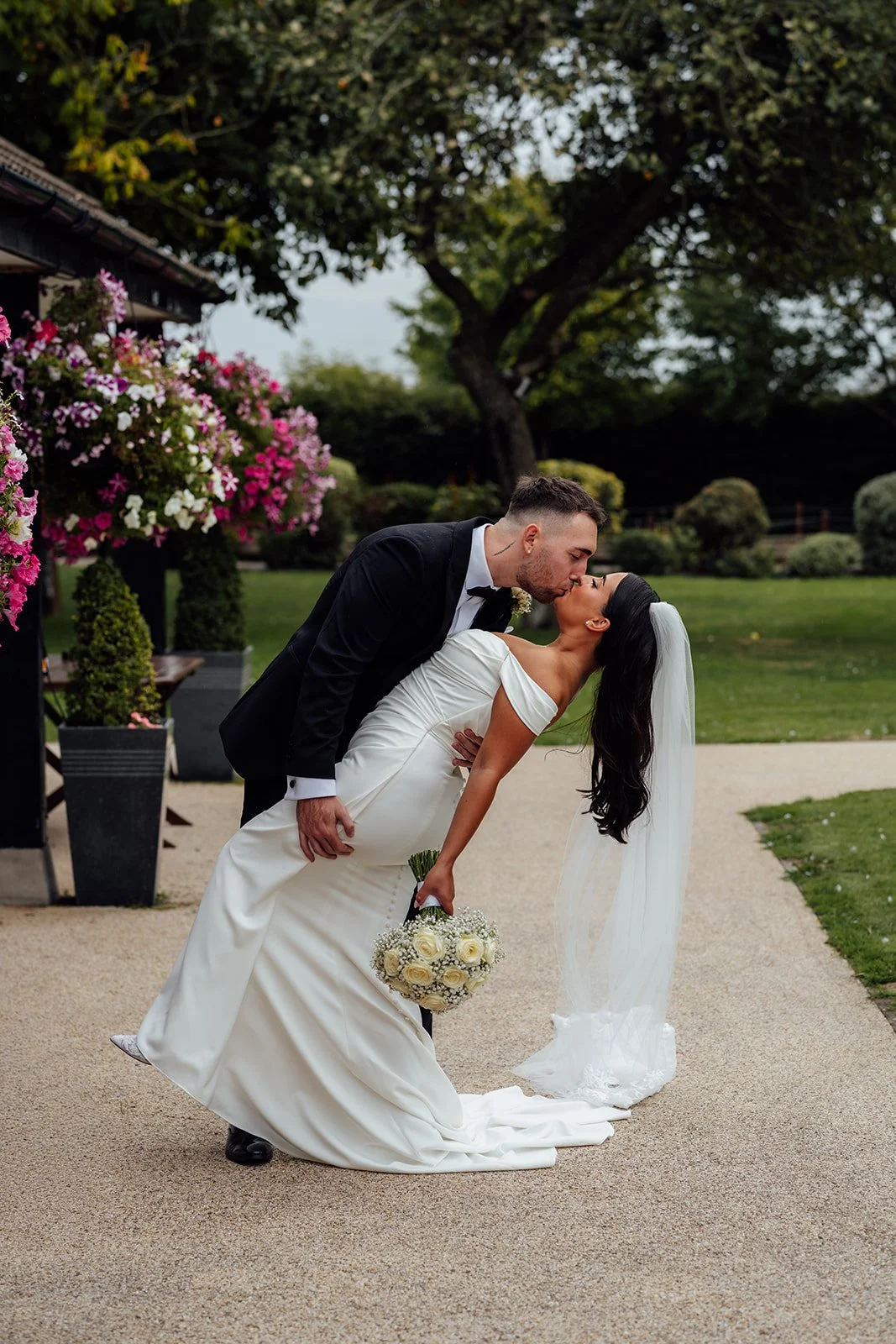Nisha and Jack share a playful dip kiss in the gardens at Batch Country House in Somerset, UK, surrounded by colourful hanging flowers and greenery as Nisha holds her round white bouquet and veil behind her, photographed by Just James Weddings.