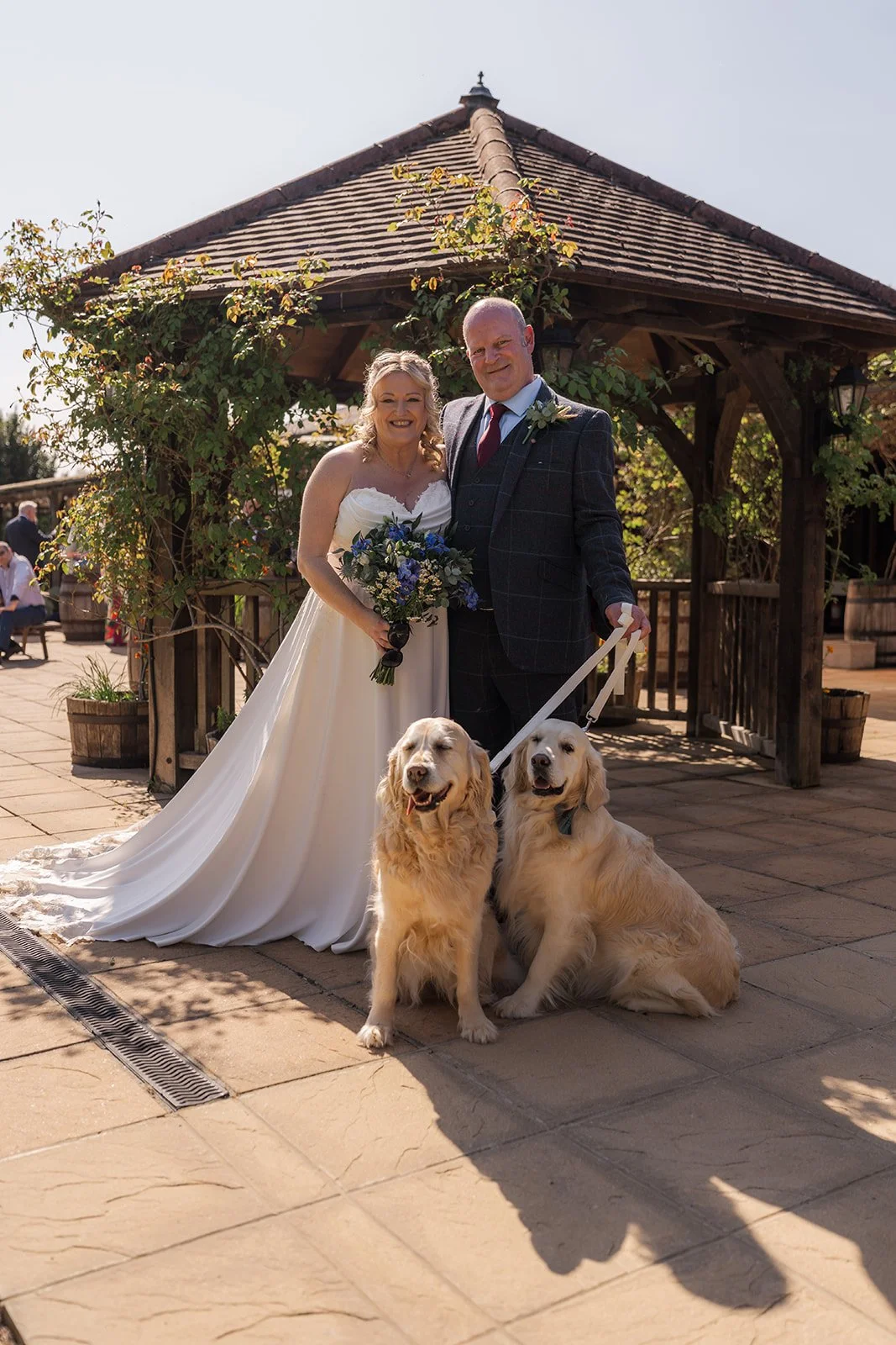Debbie and Nigel pose with their two golden retrievers outside the wooden pergola at Yarlington Barn in Somerset, creating a sunny pet-friendly wedding portrait with the rustic barn venue and gardens behind.