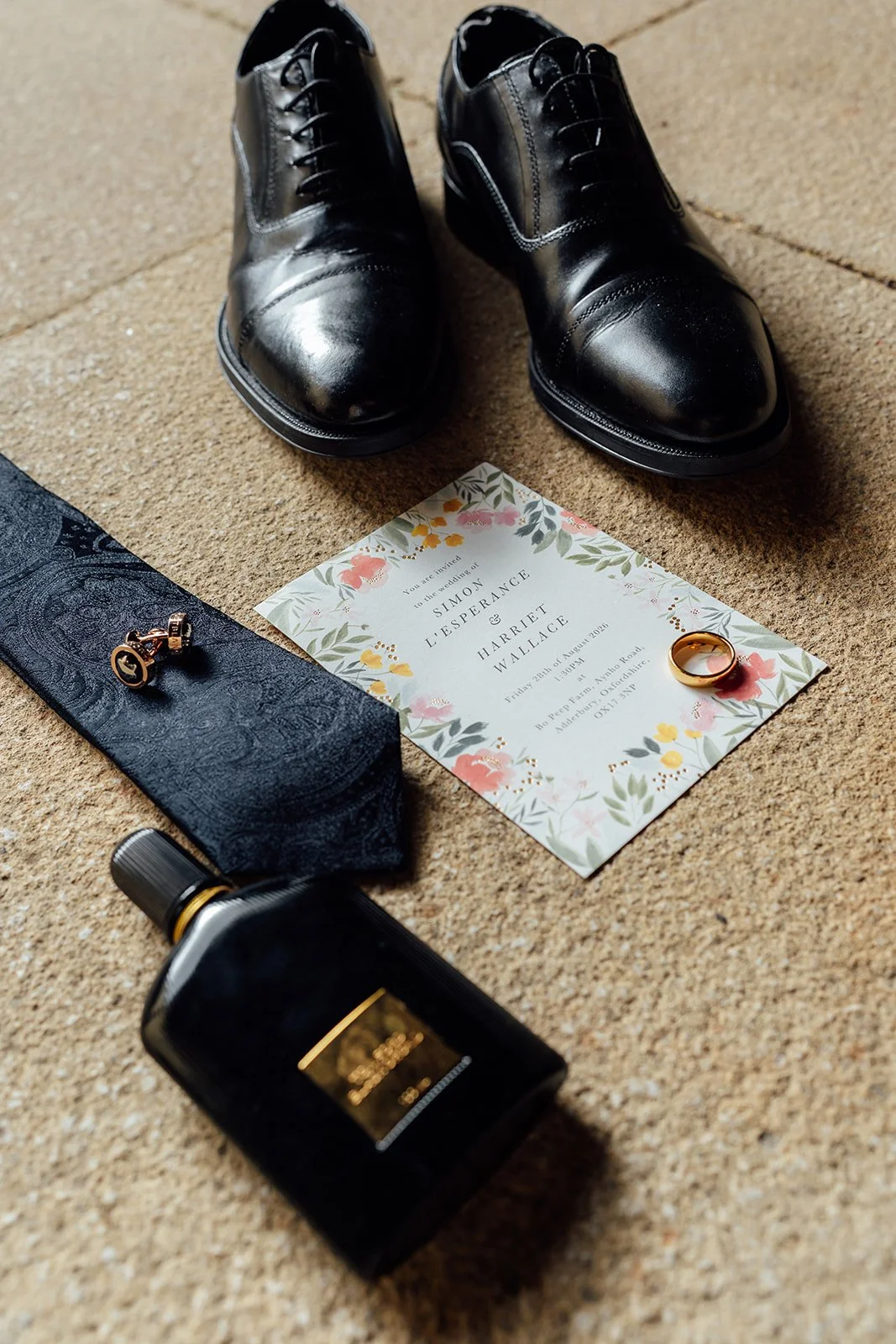 Wedding detail photo of Simon’s black shoes, patterned tie, cufflinks, fragrance, ring and floral invitation for his wedding to Harriet at Dovecote Events in Oxfordshire, photographed by Just James Weddings.