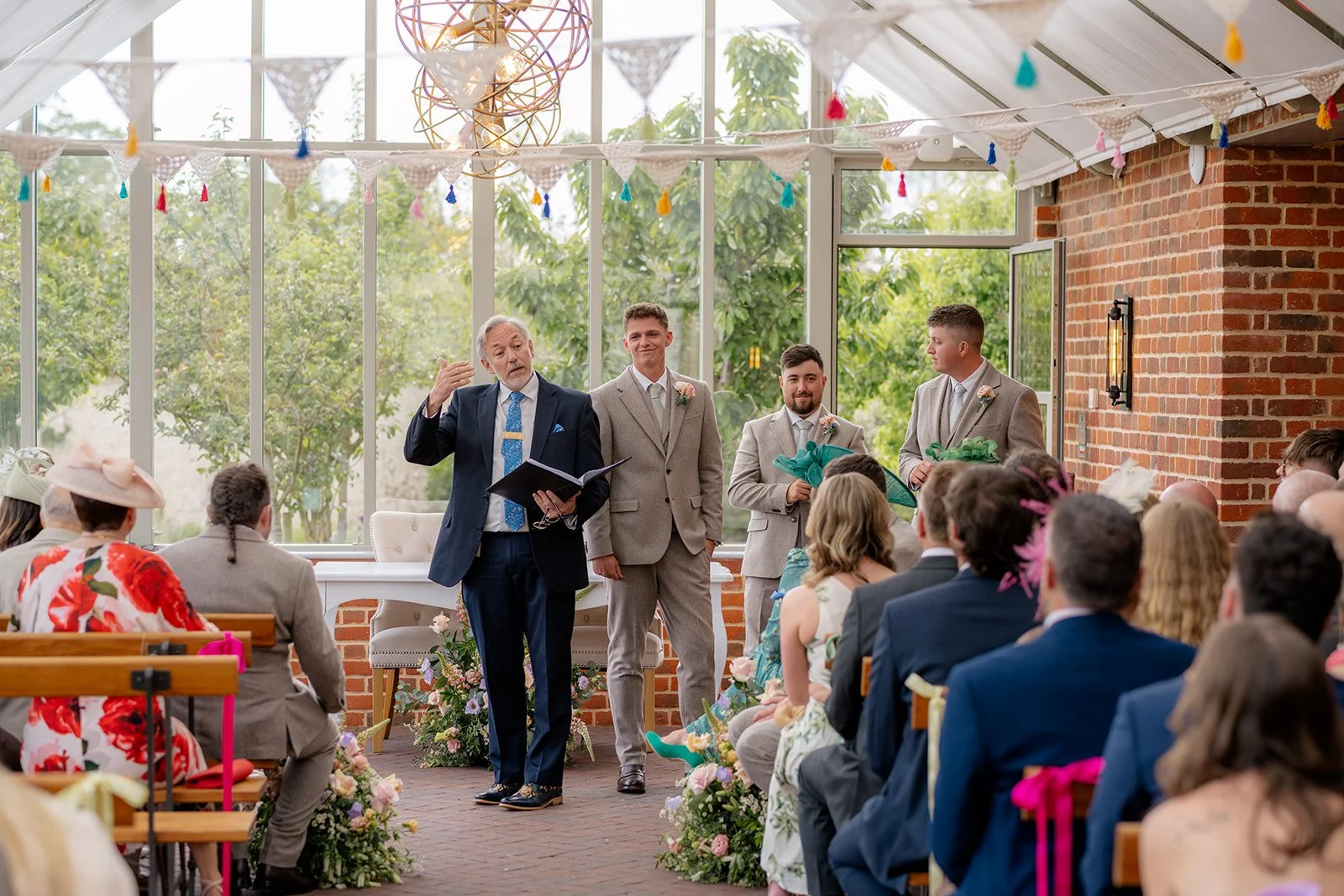 Josh standing with his groomsmen during the ceremony at Syrencot in Wiltshire, with guests seated beneath the glass-roofed venue space, photographed by Just James Weddings.