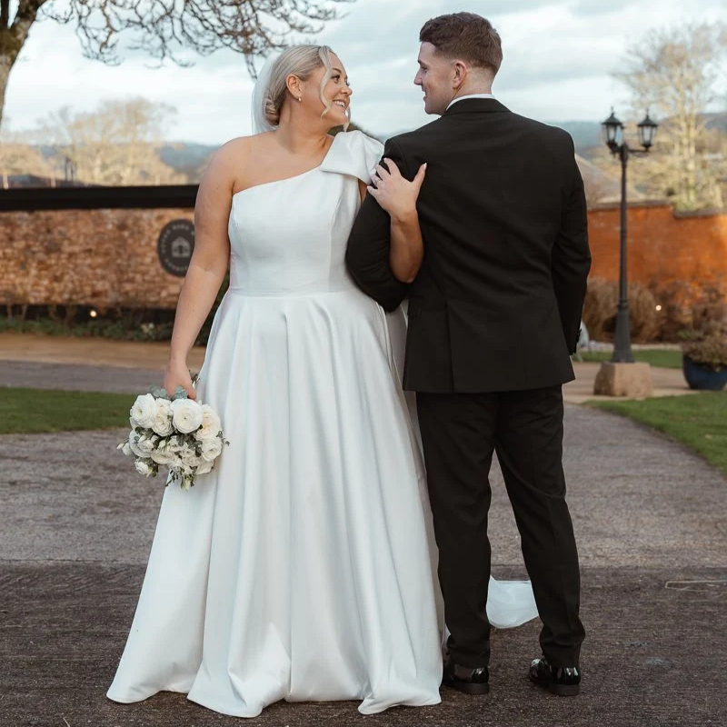 Jessie & Jay walking together outside Upton Barn & Walled Garden, devon, on their wedding day. The bride wears a one-shoulder white gown and holds a bouquet of white roses as the groom looks back at her.