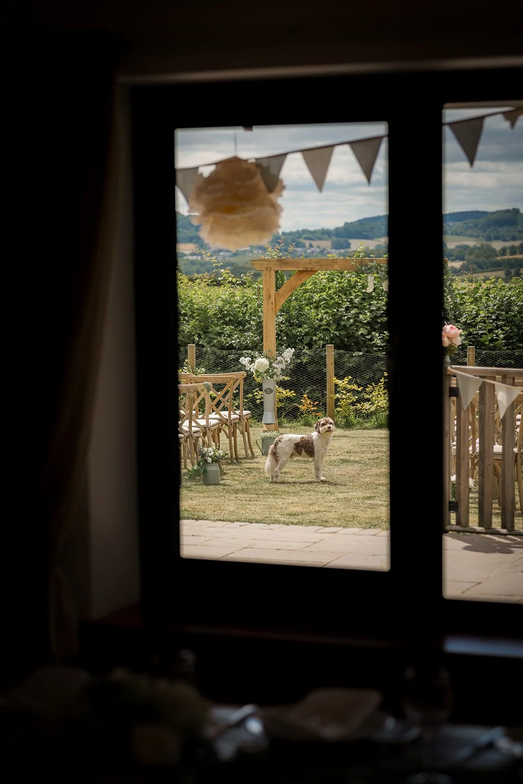 Aaron & Jack’s dog Milo stands in the outdoor ceremony space surrounded by wooden chairs, bunting and countryside views at Rock Village Hall in Worcestershire, photographed by Just James Weddings.