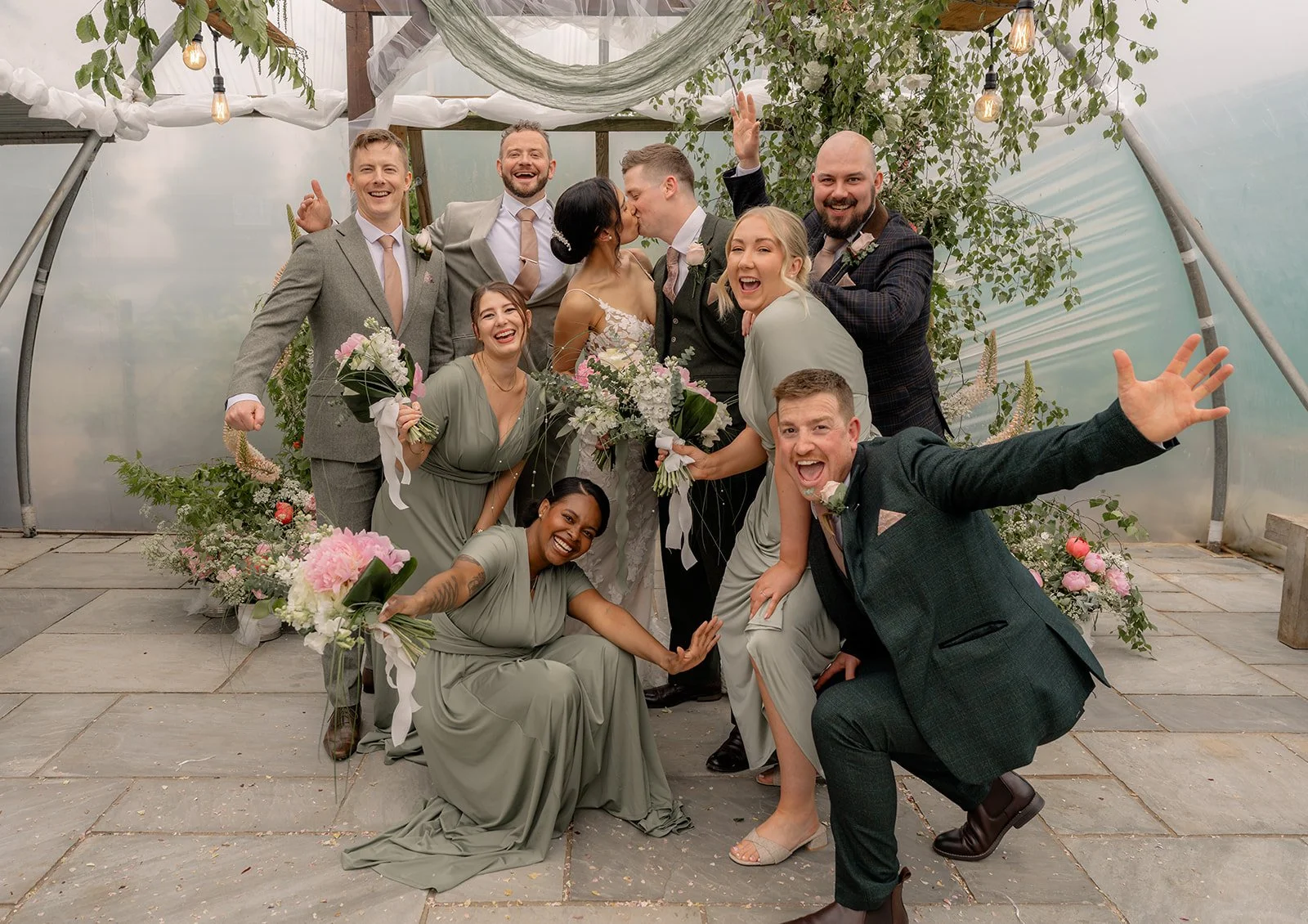 Jaye & Josh kiss in the middle of their wedding party at Farrington's Farm in Bristol, surrounded by laughing bridesmaids and groomsmen, sage dresses, green suits, pink flowers and playful poly tunnel celebration energy after the ceremony too.