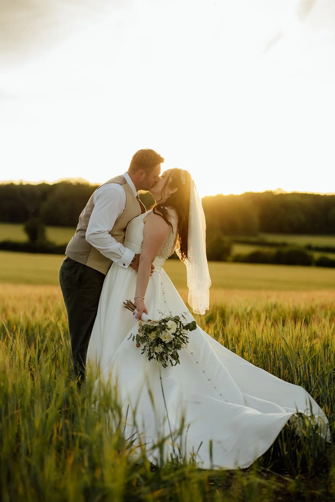 Chloe and Armani kiss in a golden-hour field beside The Barn at Upcote in Gloucestershire, with Chloe’s wedding dress and veil flowing behind them, photographed by Just James Weddings.