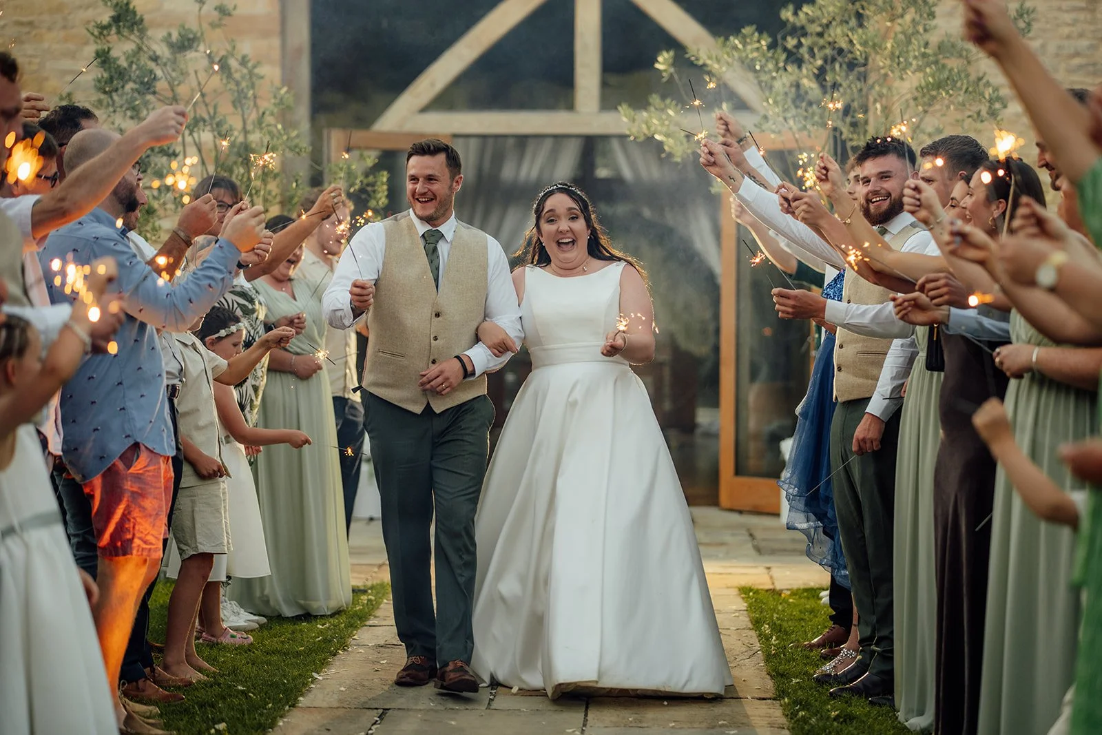 Chloe & Armani walking through a sparkler tunnel at The Barn at Upcote in the Cotswolds, surrounded by cheering guests, photographed by Just James Weddings for lively evening wedding photography.