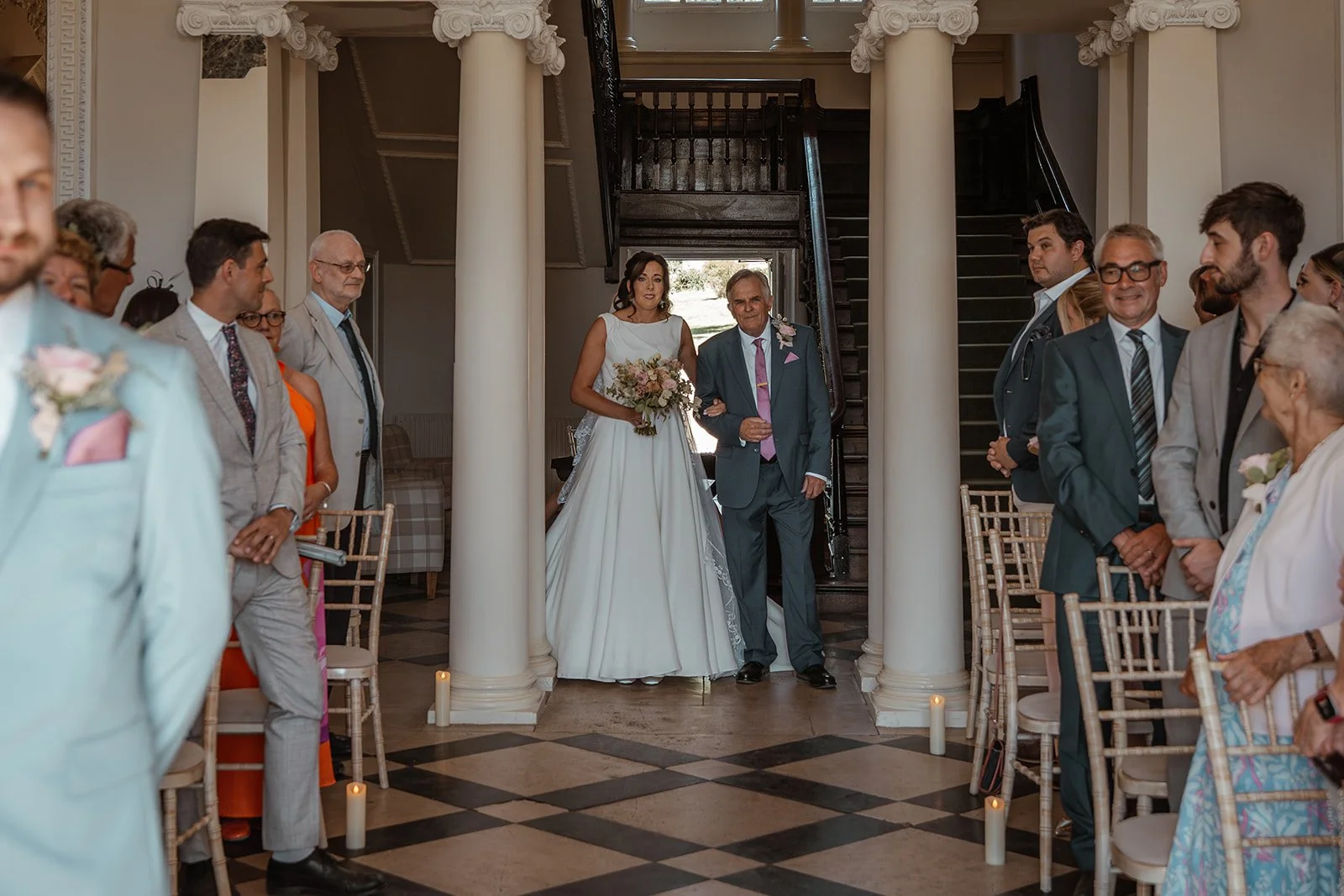 Michelle walks down the aisle with her father during her wedding ceremony at Crowcombe Court in Somerset, UK, as guests look on from either side.