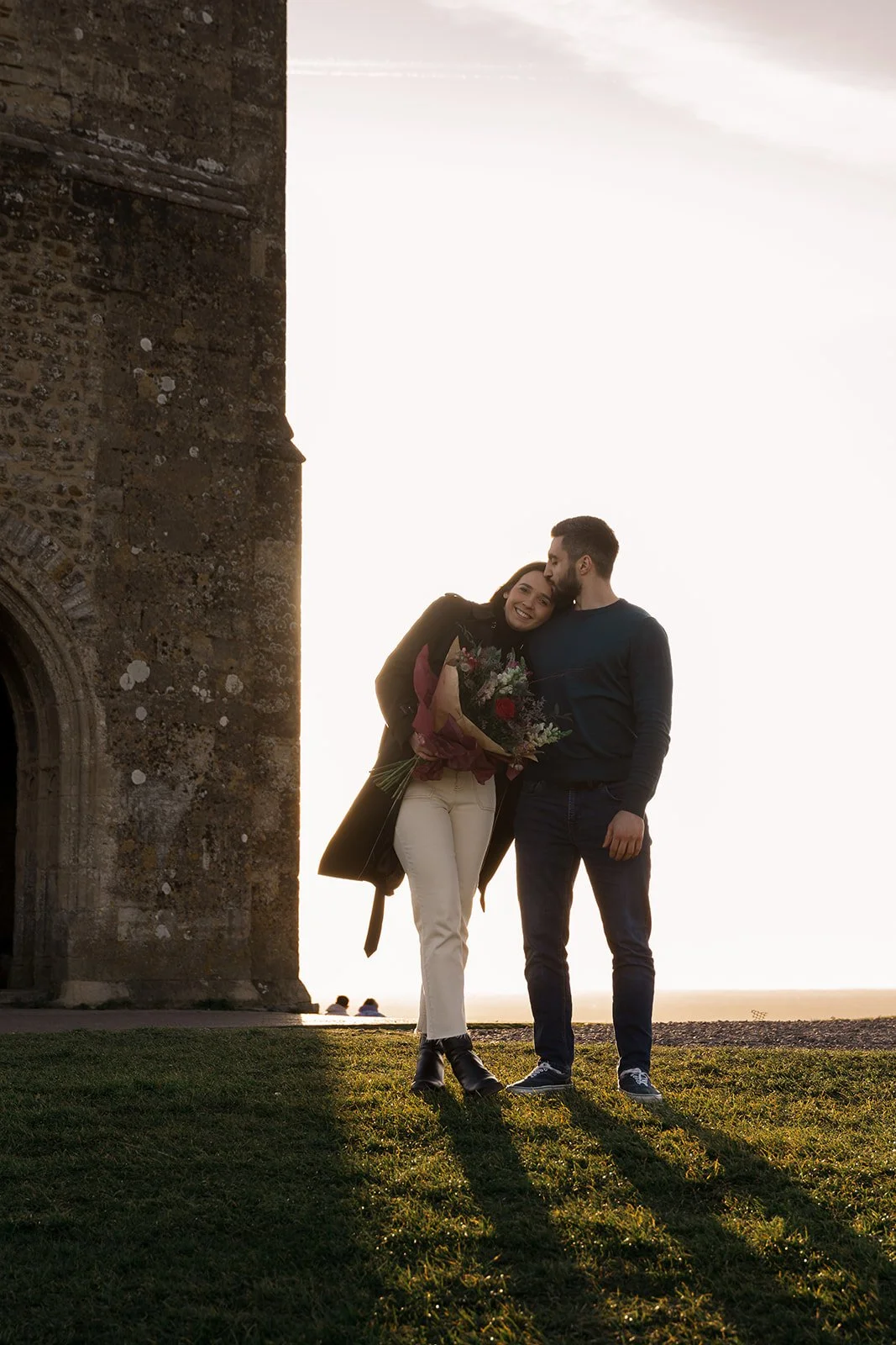 Roy and Julia standing together in warm golden evening light beside a historic stone church building in Somerset, holding a colourful bouquet after their proposal during a relaxed, natural engagement shoot photographed by Just James Weddings.