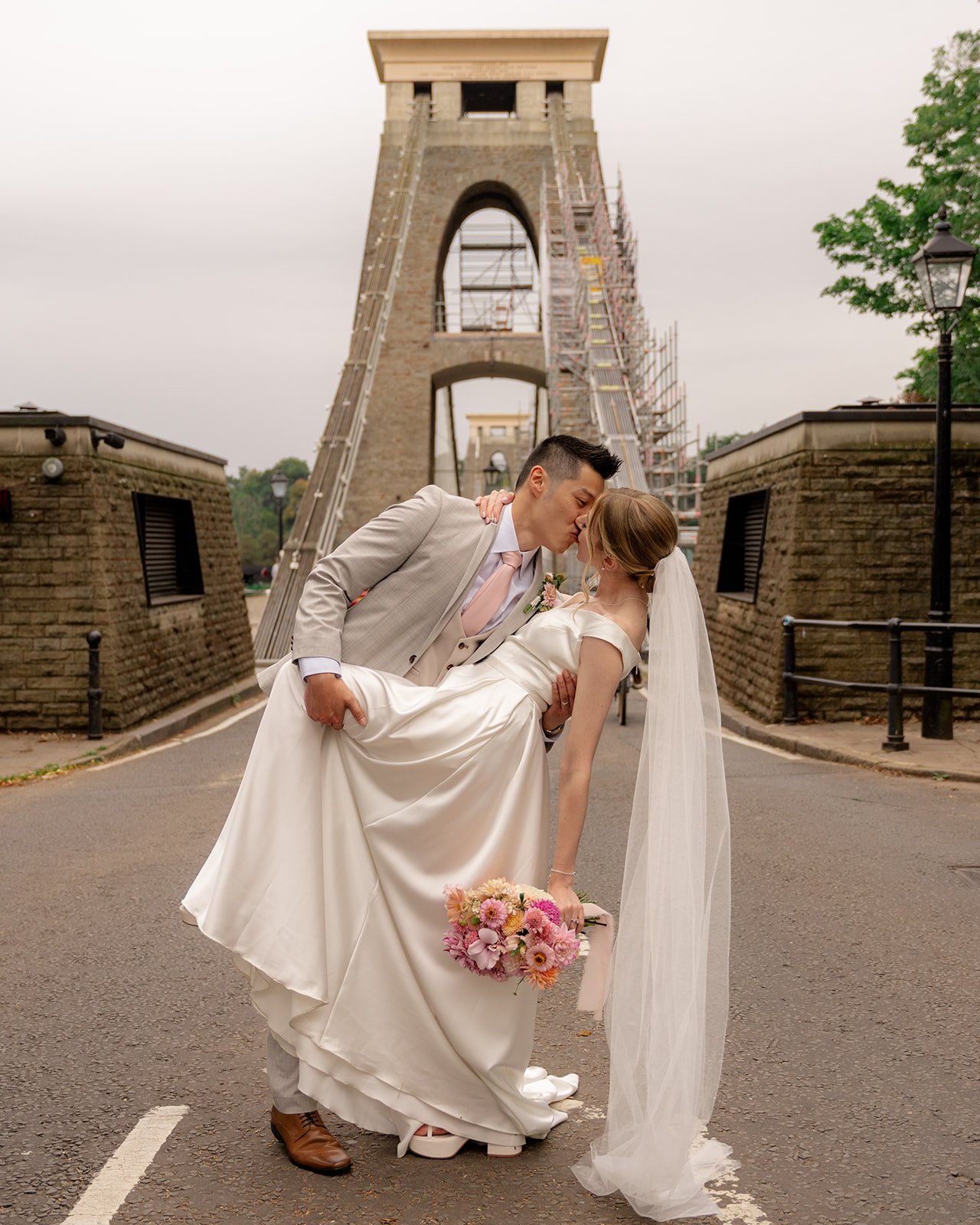 Brian & Josie kissing beneath Clifton Suspension Bridge at Clifton Observatory in Somerset, with the bride holding pink flowers, photographed by Just James Weddings.