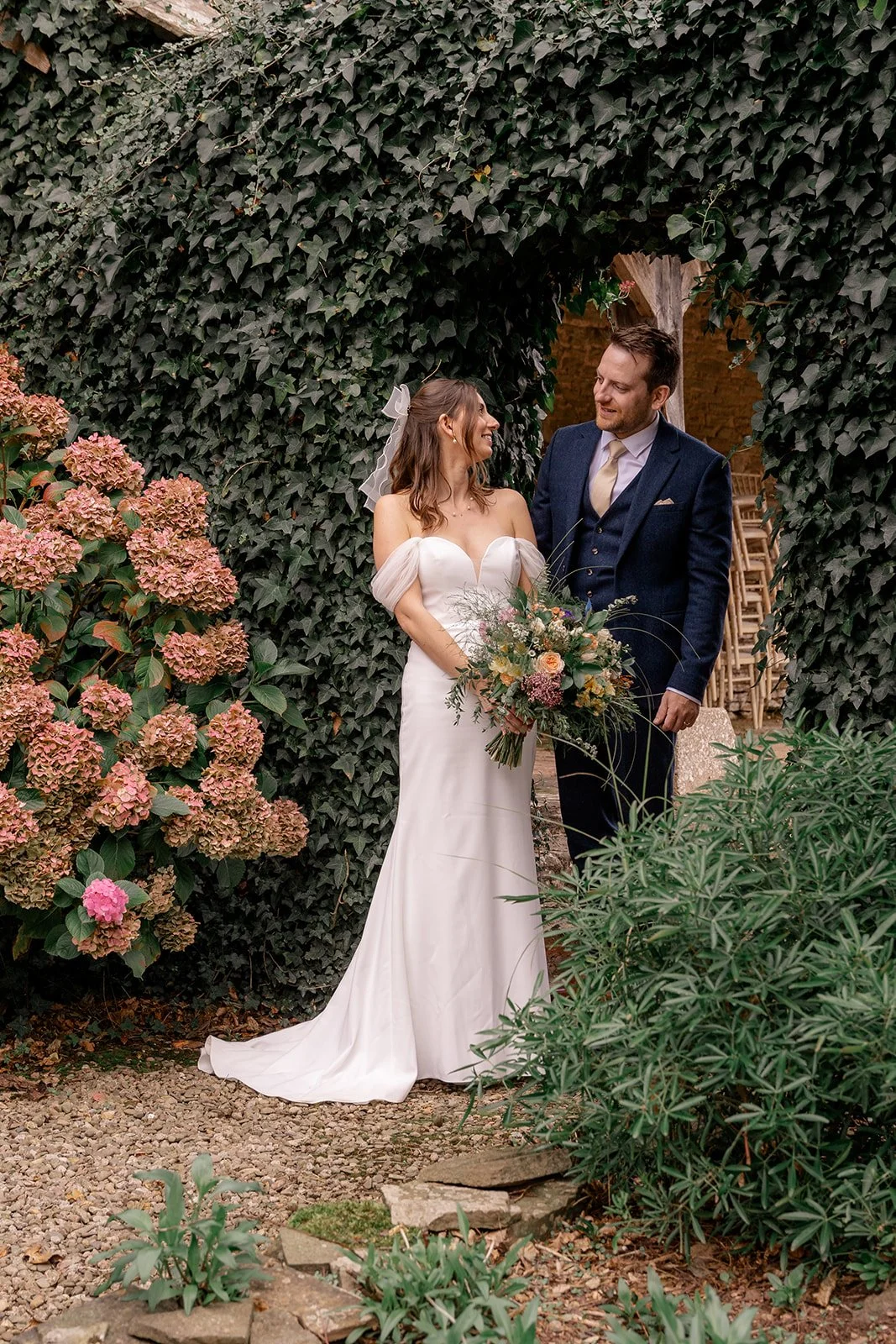 Lilly & Lloyd standing together among ivy and hydrangeas in the gardens at Broadfield Court in Herefordshire, photographed by Just James Weddings for romantic countryside wedding portraits.